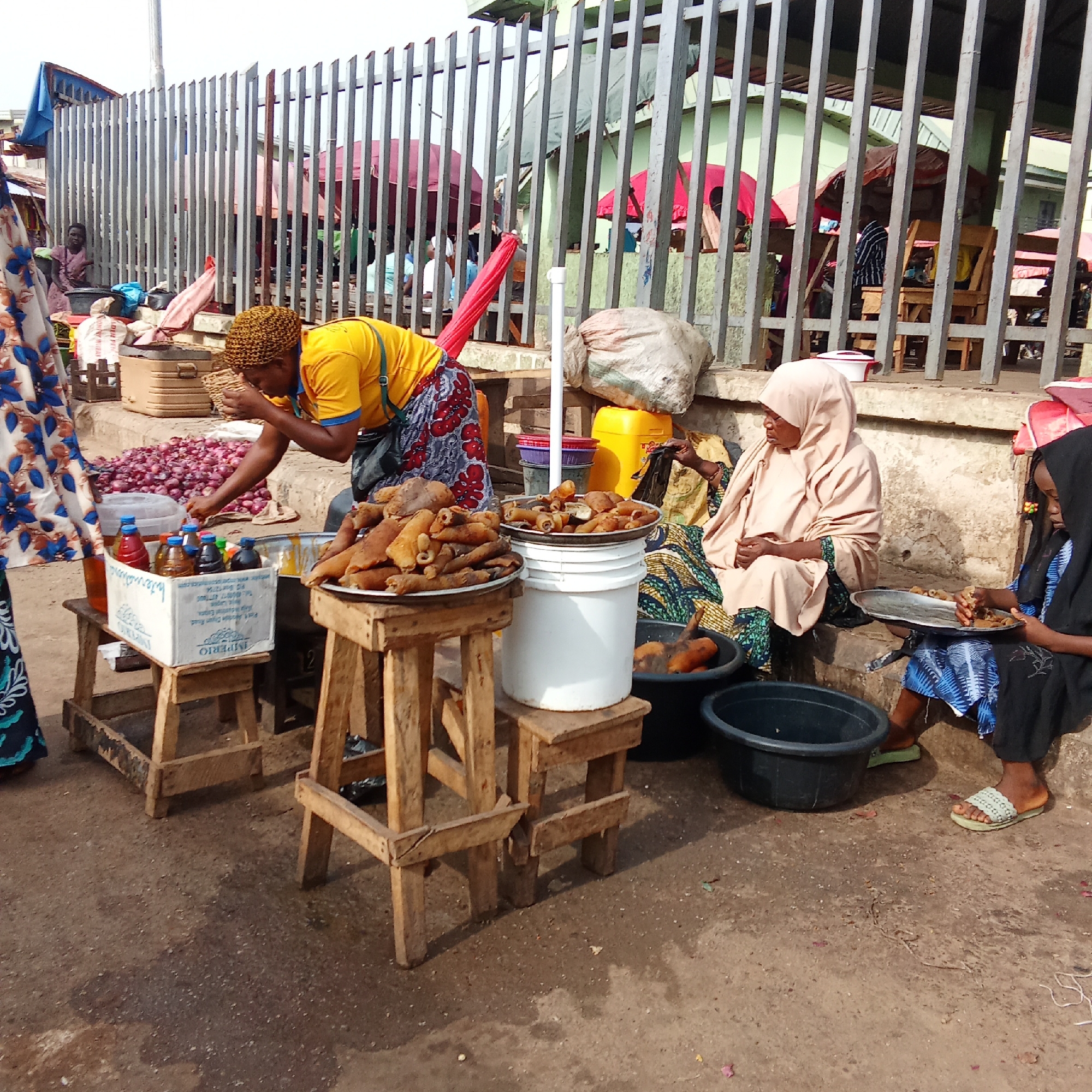 The image captures a bustling outdoor market scene in Akwanga, Nigeria. In the foreground, a woman wearing a bright yellow shirt, a patterned skirt, and a woven cap is actively engaged in preparing or arranging items for sale. She is bent over a stall where a large pile of red onions is visible.
Adjacent to her, two other individuals are seated on a low concrete ledge. An older woman, draped in a light beige hijab and a patterned green and blue fabric, sits observing the surroundings. Beside her, a young girl, wearing a dark head covering and a blue and white patterned dress, is focused on eating from a metal plate, picking at small, reddish-brown food pieces.
Prominently displayed on wooden stools are two large metal plates piled high with light brown, textured food items, which appear to be "ponmo" (cowhide), a popular Nigerian delicacy. One of these plates rests atop a white plastic bucket. A white cardboard box on another stool holds several small plastic bottles containing various colored liquids, likely beverages. The brand name "IMPERIO" is clearly visible on the box, along with some smaller, illegible text. Several black plastic tubs are scattered on the ground, some containing similar food items.
The background reveals more of the market activity, partially obscured by a tall, grey metal fence. Behind the fence, other market stalls with colorful umbrellas (red and maroon) are visible, along with other people. The ground is a mix of dirt and concrete. The diffused lighting suggests it is daytime, possibly overcast, creating soft shadows. The scene depicts a routine daily market operation.