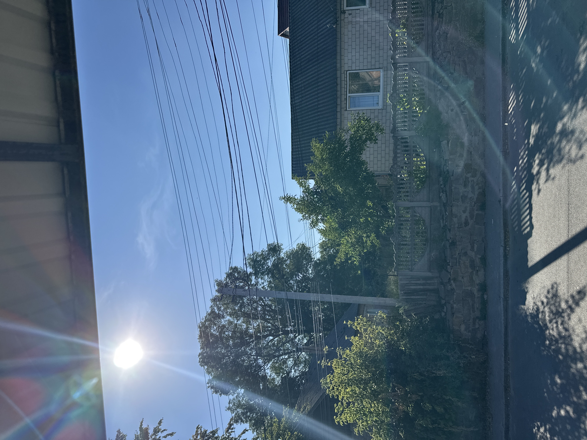 This is a photograph taken outdoors, likely during the daytime, capturing a scene in Khmilnyk, Ukraine. The image is dominated by a bright sun positioned in the upper left quadrant, casting lens flares and strong light across the frame. A clear blue sky stretches across the upper portion of the image, with several parallel power lines running horizontally. In the middle ground, a residential structure with a dark, sloped roof and light-colored brick walls is visible. The house has at least one window with a white frame. A decorative wooden fence with a scalloped top runs in front of the house, positioned on top of a low stone wall. Lush green trees and bushes are abundant throughout the middle and background, suggesting a suburban or rural setting within the city. The foreground shows a paved road or sidewalk with distinct shadows cast by overhead objects, possibly trees and a structure the photographer is standing under. The overall impression is that of a sunny day in a quiet neighborhood in Khmilnyk.