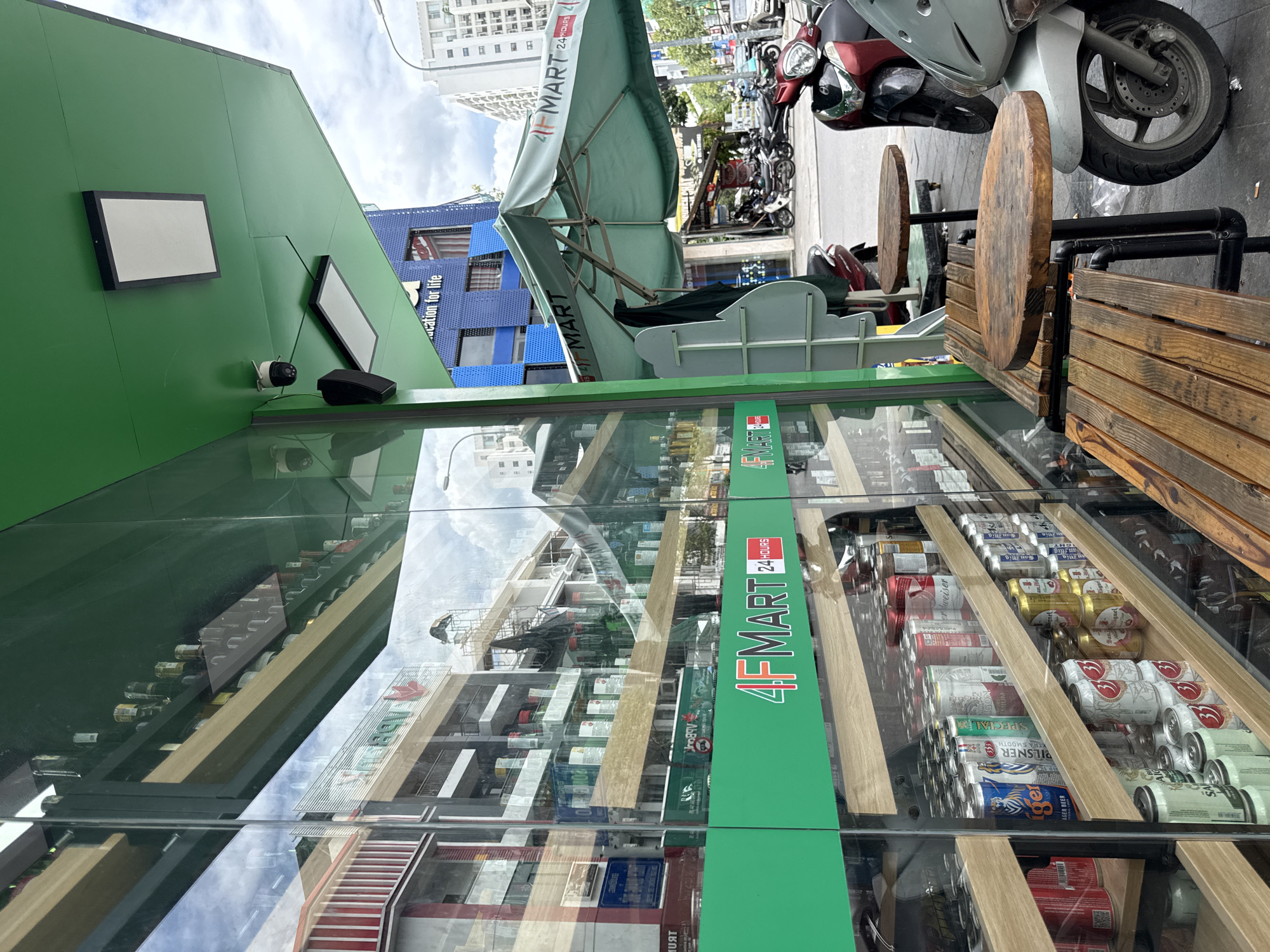 This is a street-level, eye-level shot of a 4F Mart convenience store located in Nha Trang, Vietnam. The storefront features a large glass facade reflecting the opposite side of the street, which includes buildings and cloudy skies. Inside the store, shelves are stocked with beverages, including numerous cans of beer such as Tiger, Heineken, and Budweiser, as well as bottles of what appear to be alcohol.
Outside the store, a green awning extends over a seating area with wooden benches and circular tables. A large, green umbrella with the "4F MART" logo is also displayed. Several motorcycles are parked along the sidewalk. One security camera and a speaker are visible mounted on the green overhang above the entrance. The store is identified as "4F MART 24 HOURS" with a prominent green and white sign. The scene suggests a typical urban commercial setting in Vietnam.