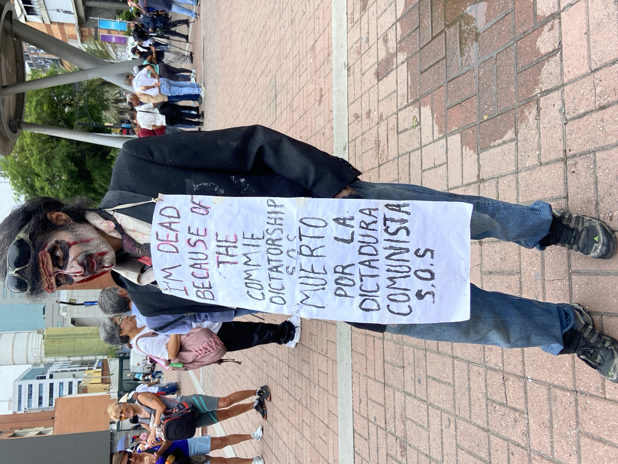 This image captures a protest or public demonstration taking place in an urban outdoor setting in Caracas, Venezuela. The ground is paved with reddish-brown bricks, some of which appear wet, suggesting recent rain.
The central subject is a person dressed in a black suit jacket, a partially visible white shirt, and blue jeans, with dark athletic shoes. Their face is covered in theatrical zombie-like makeup, featuring pale skin, sunken dark eyes, and red, bloody marks around the mouth and cheeks, conveying a gruesome appearance. Dark sunglasses rest on their head, and they have dark, disheveled hair.
Around their neck hangs a white rectangular sign, handwritten with text in both English and Spanish. The English side reads: "I'M DEAD BECAUSE OF THE COMMIE DICTATORSHIP S.O.S". The Spanish side reads: "MUERTO POR LA DICTADURA COMUNISTA S.O.S", which translates to "DEAD BECAUSE OF THE COMMUNIST DICTATORSHIP S.O.S". The overall costume and sign communicate a strong political message of distress and opposition to a communist dictatorship.
In the mid-ground and background, numerous other people are visible, some walking and others standing, suggesting they are part of the same gathering. They are dressed in casual clothing. Behind the crowd, modern buildings and green trees line the horizon, indicative of a city environment. The lighting suggests it is daytime, possibly overcast.