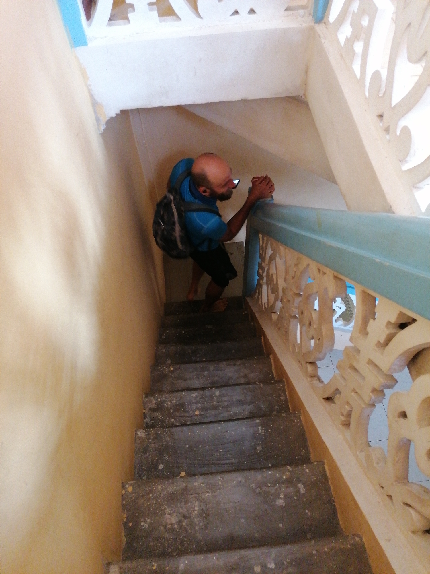 A man with a shaved head, wearing a blue t-shirt and black shorts, walks down a wooden staircase with a backpack. He is holding a smartphone in his right hand, as if taking a photo or recording a video. The staircase has a light yellow wall on the left and a decorative cream-colored railing with intricate carvings on the right. The railing is topped with a light blue trim. The man's bare feet are visible on the wooden steps. The overall environment suggests an indoor staircase within a building. The specific location within Lộc Quý, Vietnam, is not determinable from the image alone.