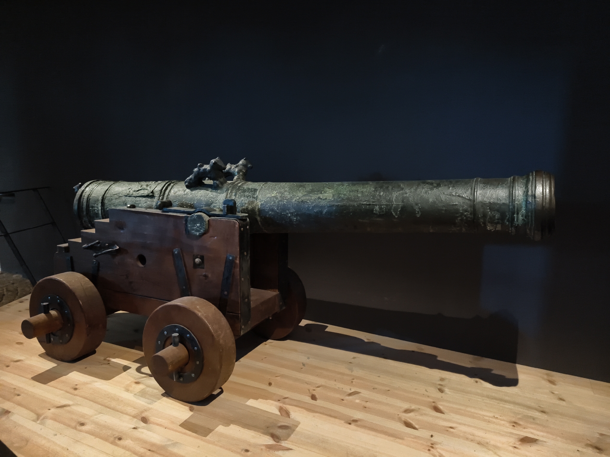 A bronze cannon with a wooden carriage sits on a wooden platform. The cannon is dark green and has intricate details. The carriage is made of light brown wood with metal accents. Two large wooden wheels are attached to the carriage. The background is a dark gray wall. The scene is lit from the right, casting a shadow of the cannon on the wall and floor. The setting is likely a museum or historical exhibit. No people are visible in the image. The object is positioned horizontally, facing right.