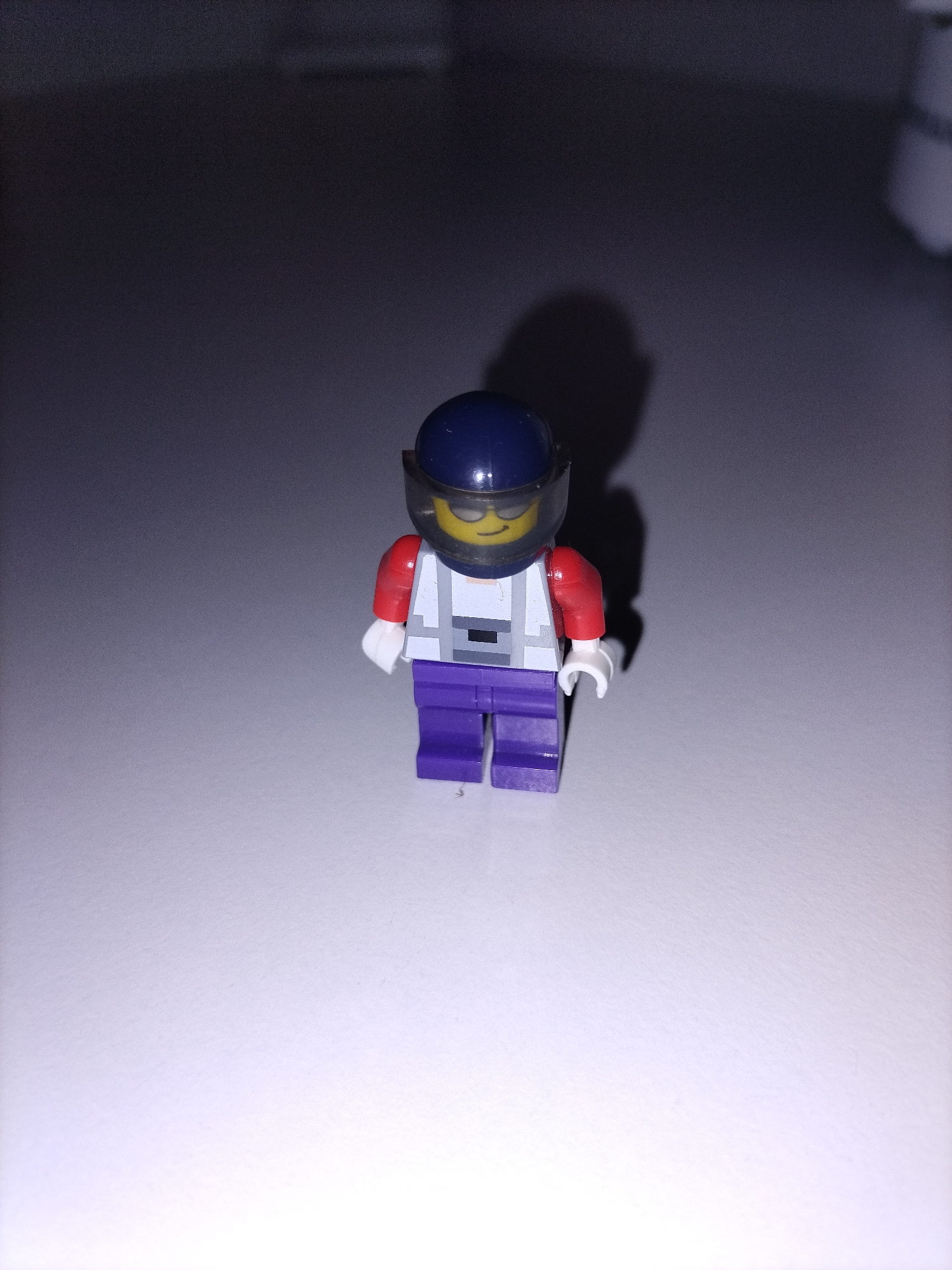 The image displays a single Lego minifigure standing upright on a flat, light-colored surface, possibly a table or floor. The background behind the minifigure is dark gray or dark purple, suggesting a shaded area or wall.
The minifigure is wearing a dark blue helmet with a translucent yellow visor pulled down, covering its face, which appears to be the classic Lego smiley design. Its torso is white, detailed with light gray and dark gray patterns, resembling a racing suit or vest. The arms are red with white hands, and the legs are purple.
The lighting originates from the front, casting a distinct, dark shadow of the minifigure behind it and slightly to its right. The surface in the foreground is brightly lit, while the overall image shows some digital noise or grain. There are no activities taking place other than the minifigure standing. No specific location cues are visible beyond an indoor setting, and there is no discernible text.