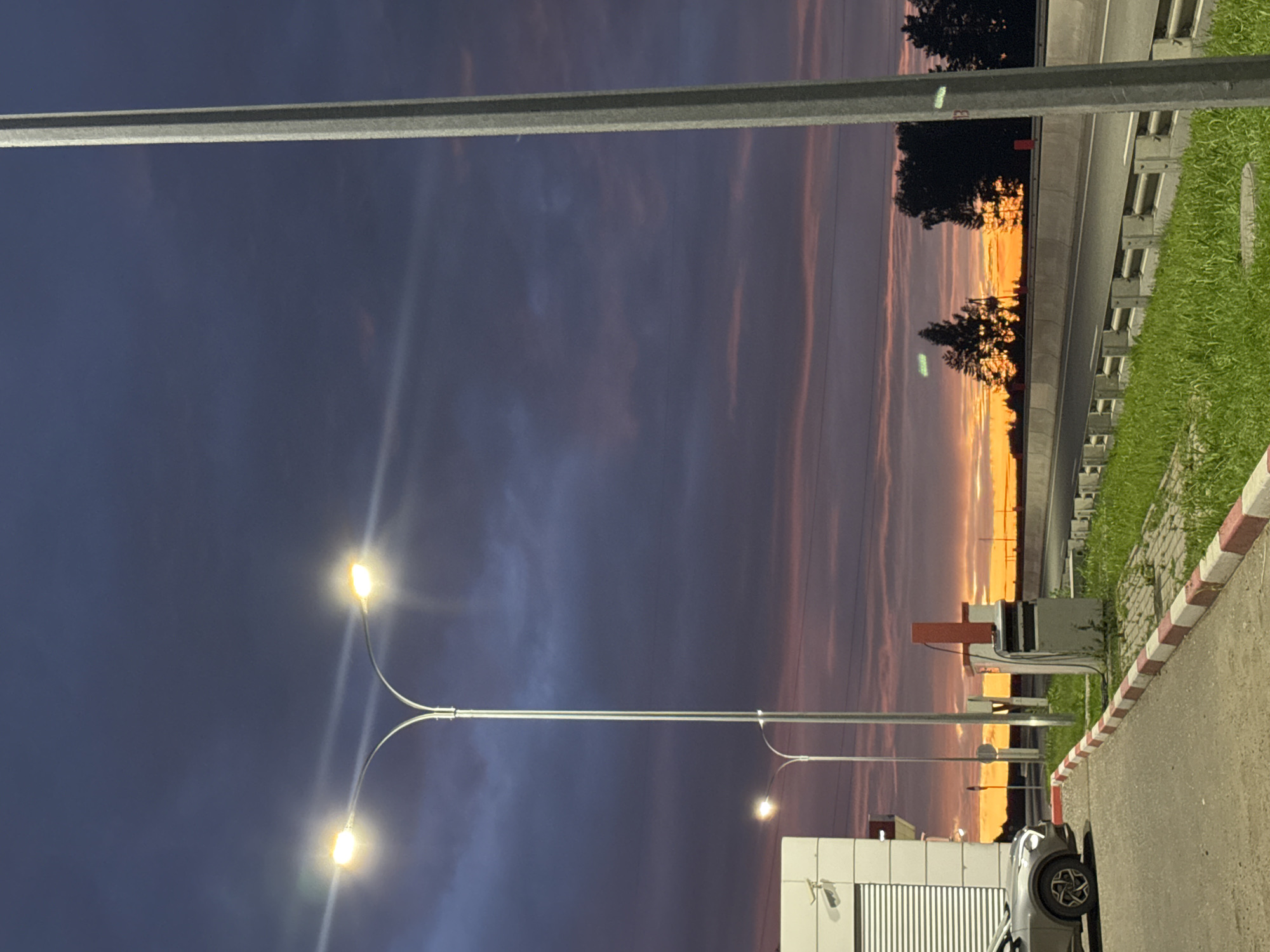 This media file depicts an outdoor scene during twilight, likely in Можга, Russia. The sky is filled with dramatic clouds tinged with orange and purple hues, suggesting a sunset or sunrise. Tall streetlights with illuminated bulbs are prominent in the foreground, casting a glow against the darkening sky. A road or pathway is visible, bordered by a curb with red and white markings. To the right, a patch of green grass and a concrete barrier with regularly spaced indentations are present, possibly indicating a roadside or park area. In the background, silhouetted trees are visible against the vibrant sky, and the lights of distant structures can be seen. A portion of a building with a ribbed facade and a security camera is in the lower right corner, along with the rear of a vehicle. The overall atmosphere is calm and serene, with the vibrant colors of the sky being a focal point. The time of day is clearly twilight, given the illumination of the streetlights and the colors in the sky. There is no visible text in the media file.