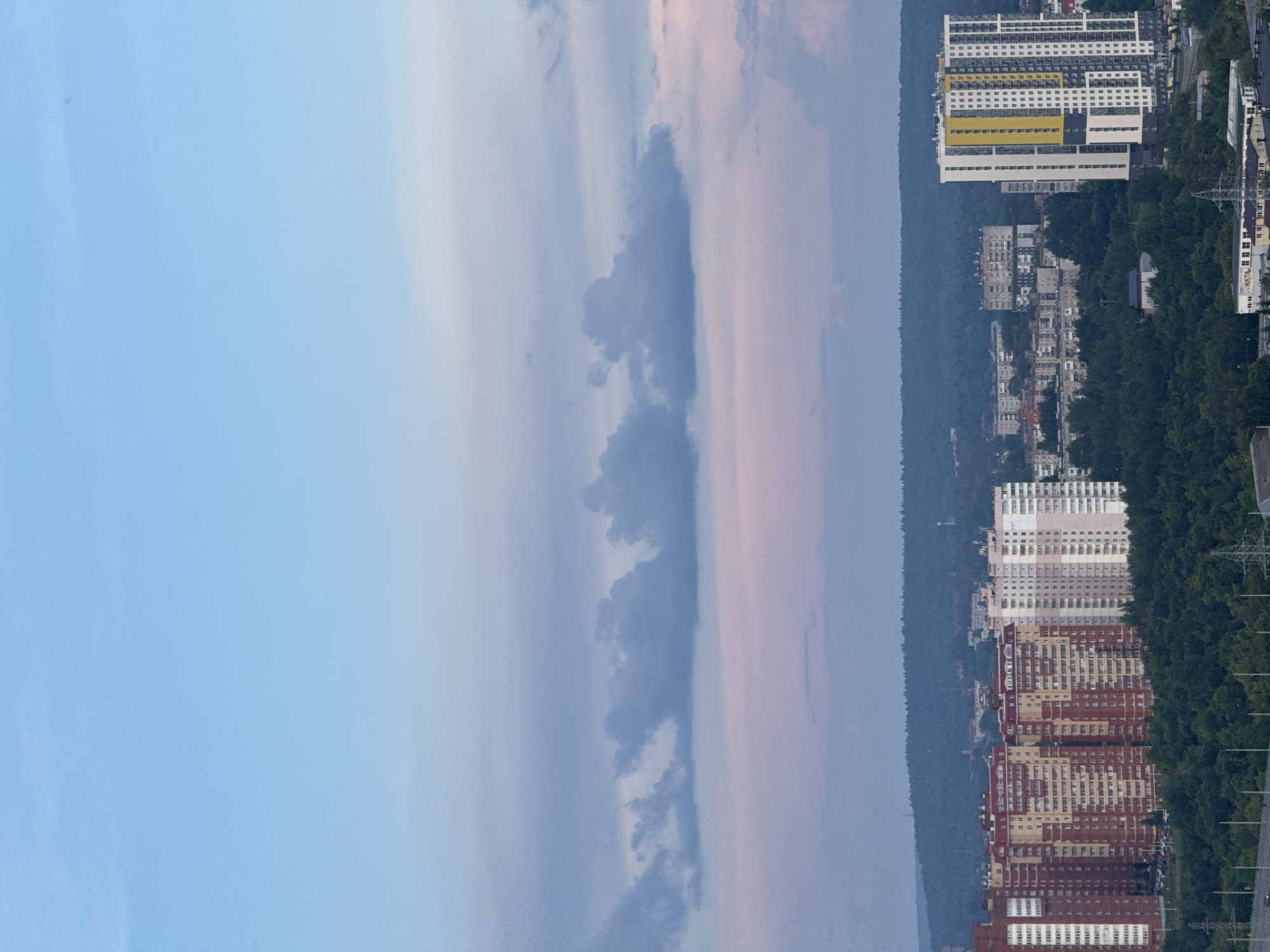 This is a vertical, high-angle, exterior photograph of a residential area with many apartment buildings. The buildings are tall and vary in color, with some featuring red, yellow, and white facades. They are situated next to a dense forest. The sky above is a mix of pale blue and pink hues, with some clouds present. A power line tower and road are visible to the right of the apartment buildings, suggesting an urban or suburban setting. The overall lighting suggests it might be during sunset or sunrise. There is no visible text in the image.
