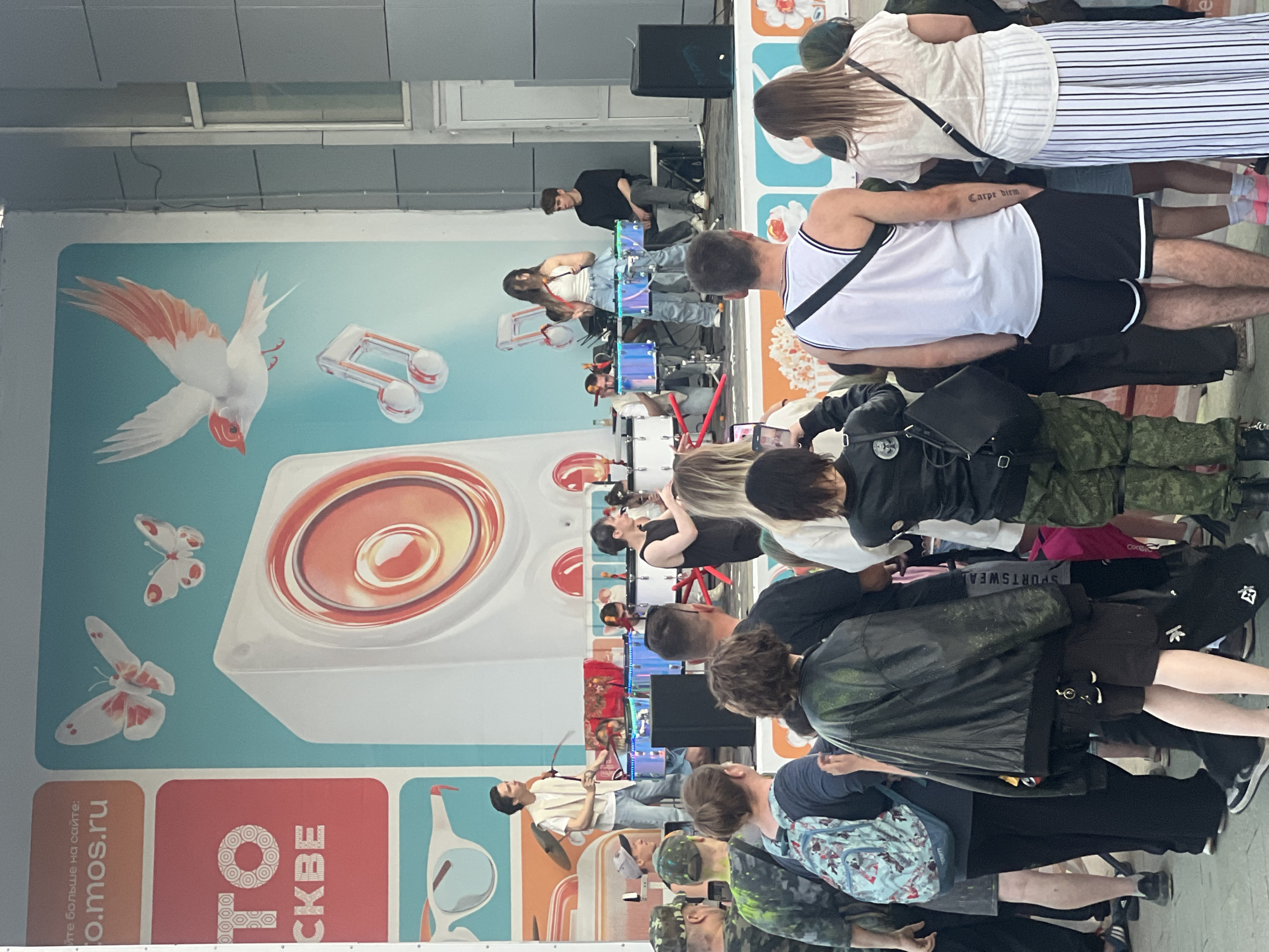 The image depicts an outdoor performance scene in Moscow, Russia, likely in a public space or entertainment venue. A stage is set up with a large backdrop featuring graphics of musical notes, butterflies, and a stylized bird. In the center of the backdrop is a large image of a white speaker.
On stage, a musical group appears to be performing. A male performer in a light-colored vest and jeans stands at the front, holding drumsticks and facing towards a set of drums. Beside him, a female performer in a white crop top and jeans is also positioned near drums. Other performers can be seen behind them, possibly playing drums or other instruments, with some seated. The drums themselves appear to be illuminated with blue light. A male performer in a black t-shirt is seated on the right side of the stage.
In front of the stage, a crowd of spectators is gathered. They are standing and watching the performance. Several people in the foreground are facing the stage, with some holding up phones, likely to take pictures or videos. The spectators are diverse in age and attire. The overall atmosphere appears to be lively and engaged, with elements of urban street culture suggested by the backdrop and the clothing of some of the performers and audience members. The lighting indicates it is daytime.