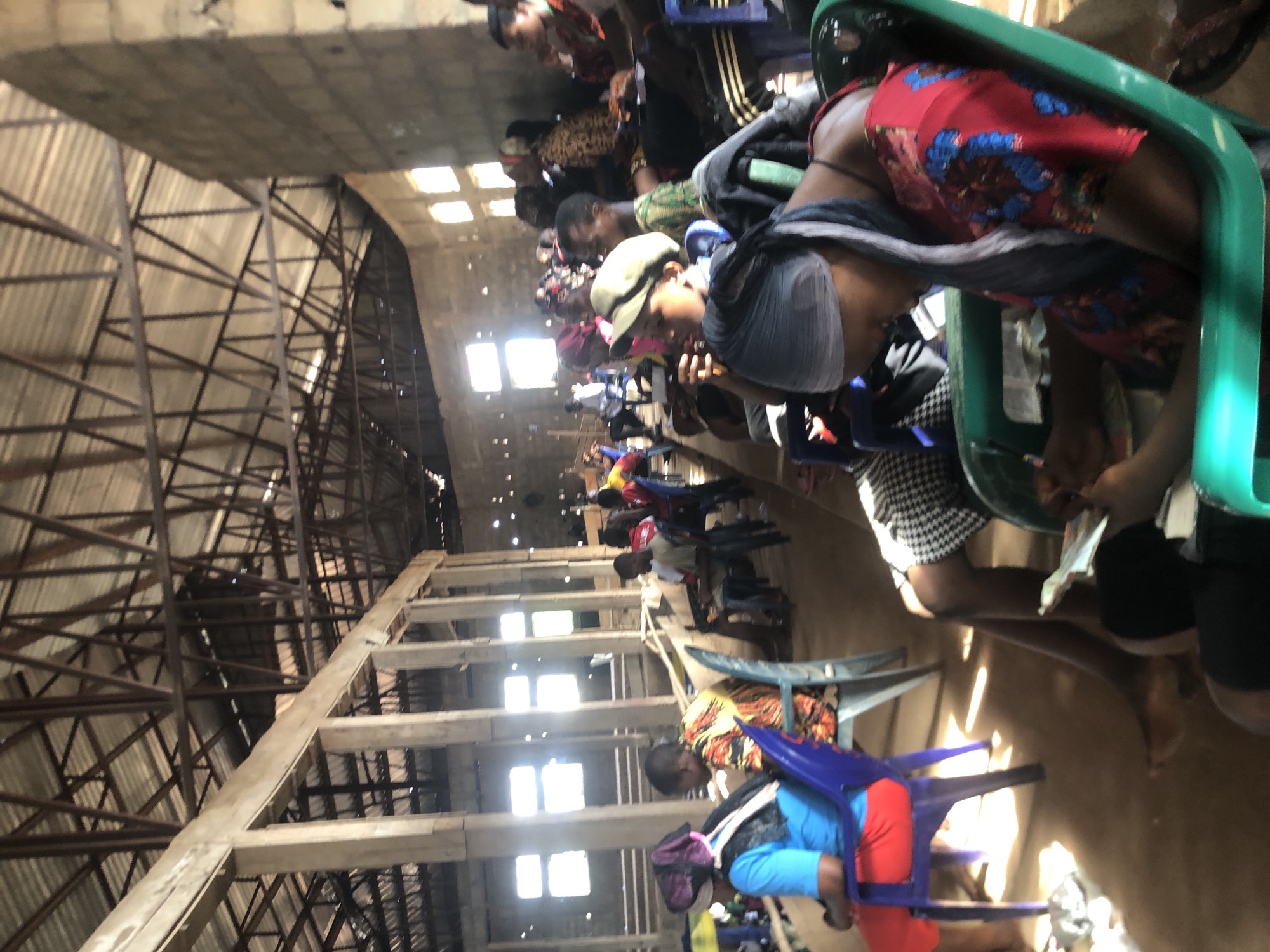 In Nteje, Nigeria, a large indoor space with a high, exposed roof structure of steel beams and support columns houses numerous people seated in plastic chairs arranged in rows. Sunlight streams in through openings in the roof, illuminating the individuals as they are bent over, likely writing or reading, presumably taking a test or examination. The floor appears to be bare and unmarked. The scene suggests a temporary testing center.