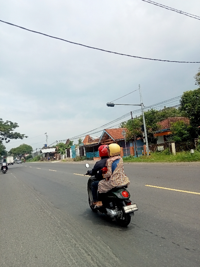 The image shows a man and a woman riding a motorcycle on a paved road in Kecamatan Margorejo, Indonesia. The man is wearing a red helmet and a blue shirt, and the woman is wearing a yellow helmet and a striped shirt. They are riding on a green scooter, and the woman is holding onto the man. There are some houses on the right side of the road, and a couple of cars in the distance. The weather is cloudy, and the road is straight with a faint yellow stripe down the middle. The location is near the coordinates -6.787 latitude and 110.981 longitude, in a residential area. The motorcycle has a license plate that reads "4265 PU". The motorcycle is facing forward, and the man and woman are looking straight ahead.