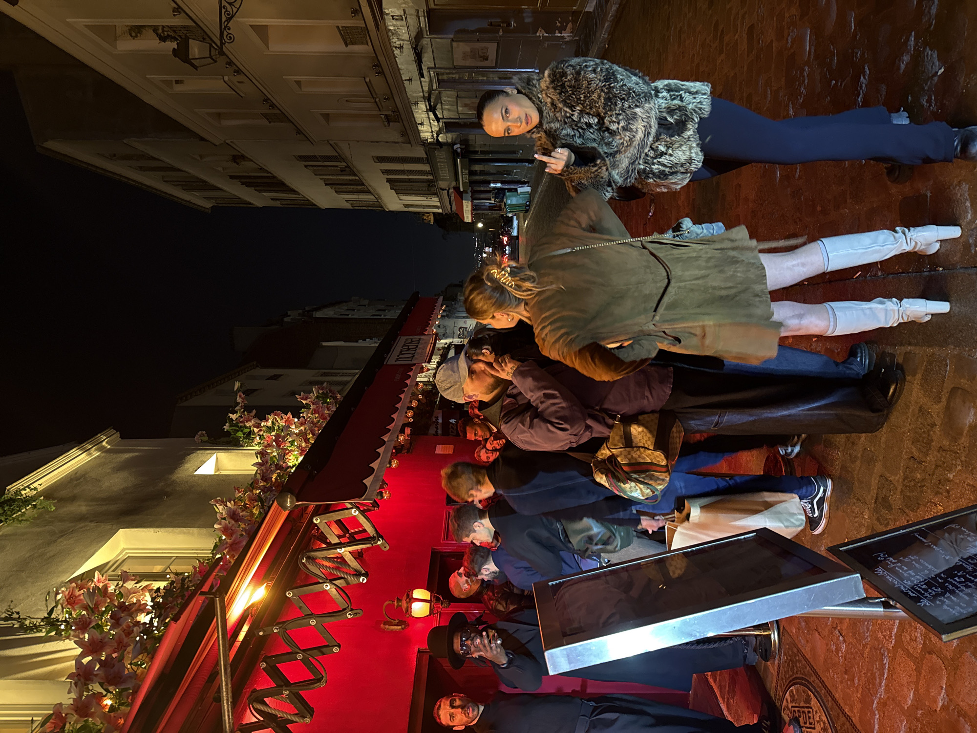 The image depicts a nighttime street scene in Paris, France. A group of people are gathered in front of a red storefront decorated with flowers. The storefront has an awning supported by a black, extendable scissor arm. The people are dressed in casual attire, some wearing coats and hats. One person is standing on the sidewalk, wearing a fur coat and giving the middle finger. The buildings across the street are visible, with windows and decorative architectural details. The ground appears to be wet, reflecting the streetlights. There are metal stands displaying framed images on the right of the photo.