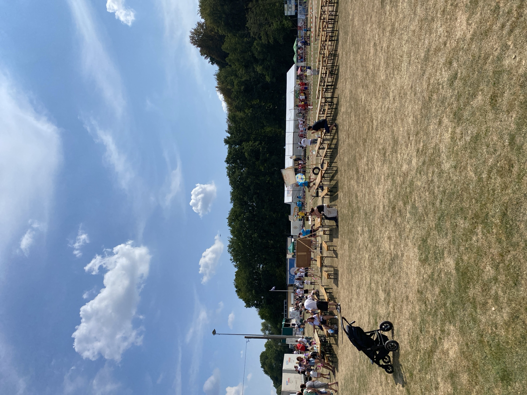 This image depicts an outdoor event, likely a festival or gathering, taking place on a dry, grassy field in Ingelbach, Germany. The sky is bright blue with scattered white clouds. A line of trees forms the background, with some tents and temporary structures visible among them.
In the foreground, a black stroller is parked on the grass. The middle ground is populated by numerous wooden picnic tables and benches, some of which are occupied by people. A crowd of individuals, dressed in casual summer attire, are scattered throughout the area, some standing and talking, others sitting at the tables. Several food stalls or vendor tents are visible further back. A tall light pole stands on the left side of the image. The overall atmosphere appears relaxed and social, suggesting a daytime event.