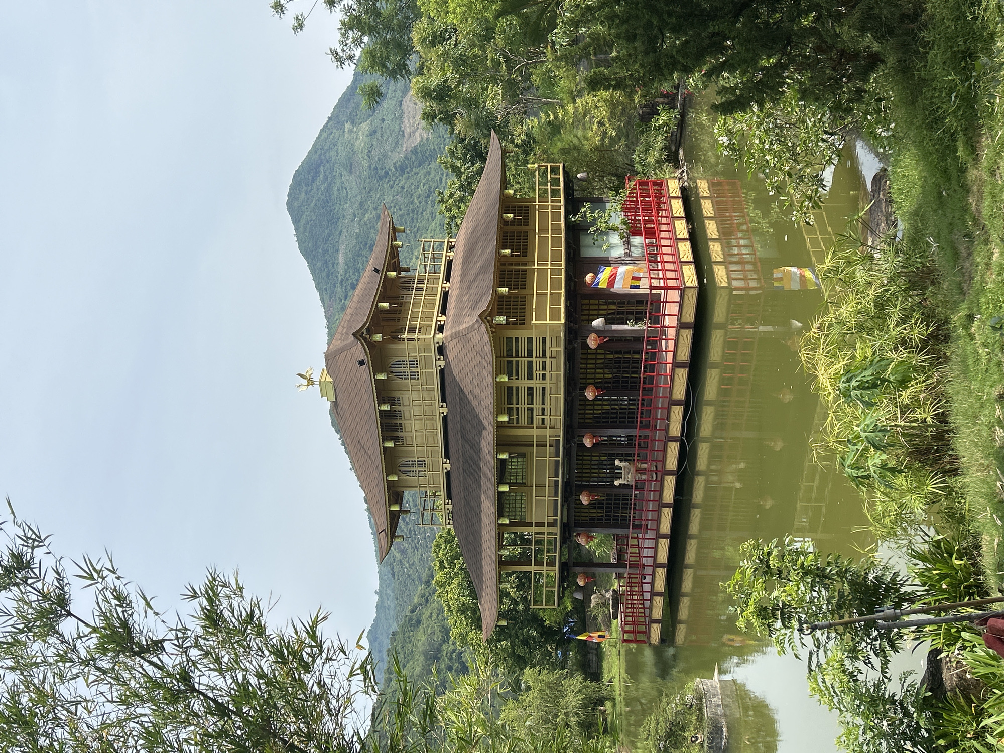 An ornate, multi-tiered golden building with a dark brown, pagoda-style roof sits on a platform above a tranquil body of water. The building features detailed yellow railings on its balconies and is surrounded by lush green trees and foliage. A backdrop of verdant, rolling hills completes the serene landscape. A Buddhist flag is visible on the left side of the platform, and red lanterns hang from the eaves of the structure. The building appears to be a Buddhist temple or pavilion, likely located in a natural, park-like setting. The water reflects the building and surrounding greenery. The scene suggests a peaceful and aesthetically pleasing environment, characteristic of temples or gardens in Vietnam.