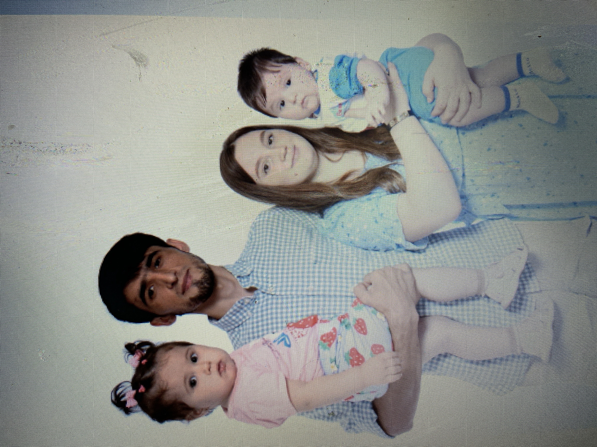 A family of four, consisting of two adults and two young children, poses for a photograph against a plain, light-colored backdrop. The father, on the left, is holding a baby girl in his arms. He has dark hair and a beard and is wearing a blue and white checkered shirt. The baby girl has dark hair styled with pink bows and is wearing a pink top and strawberry-patterned bottoms. To the right of the father, the mother is holding another baby, this time a boy. She has long brown hair and is wearing a light blue dress. The baby boy is wearing a blue outfit with white socks. The overall lighting is bright, suggesting an indoor setting, likely a photography studio. The image is a still photograph, capturing a posed family portrait. There are no discernible environmental features indicating a specific location within the city of Marghedar, Tajikistan.