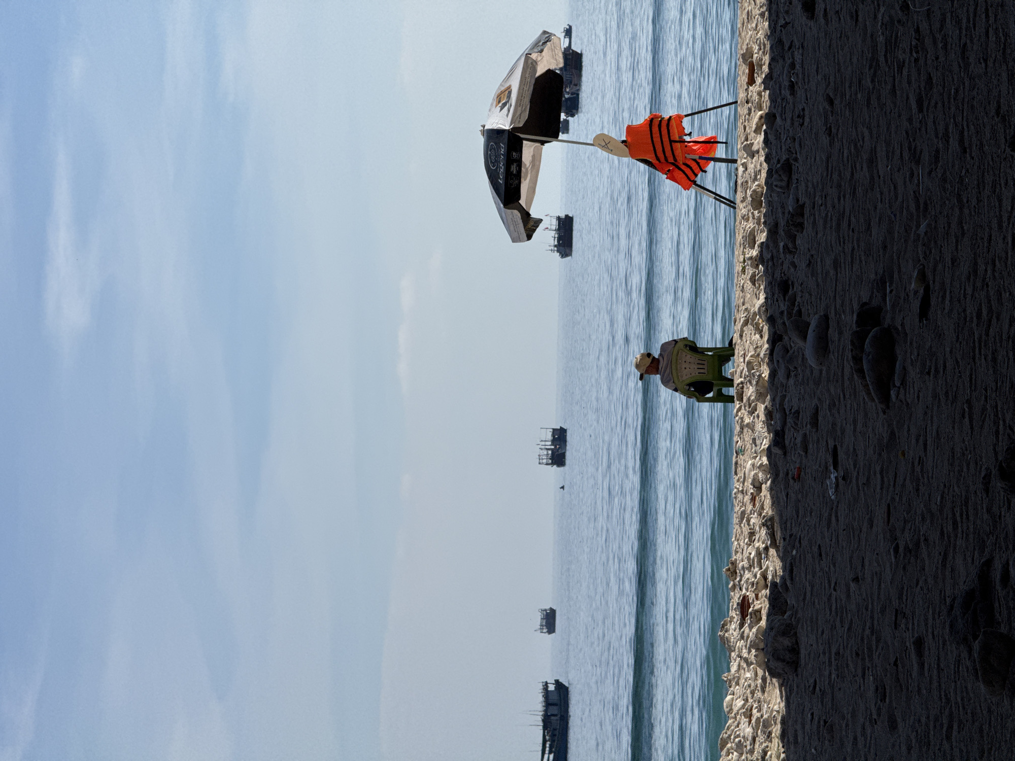The image depicts a tranquil beach scene on Lý Sơn Island, Vietnam. In the foreground, the dark sand of the beach is partially shadowed, with scattered rocks and debris visible. The middle ground features the turquoise ocean, with gentle waves lapping at the shore. Several fishing boats are scattered across the water, some appearing to be anchored or moving slowly.
On the beach, two figures are seated. To the left, a person wearing a cap is seated in a green plastic chair, facing the ocean. To the right, a black and white umbrella with the brand "Legend" is propped up, and an orange life vest is draped over a stand beneath it. The sky above is a pale blue with wispy white clouds. The overall atmosphere is one of quietude and calm, with the natural beauty of the island's coastline taking center stage.