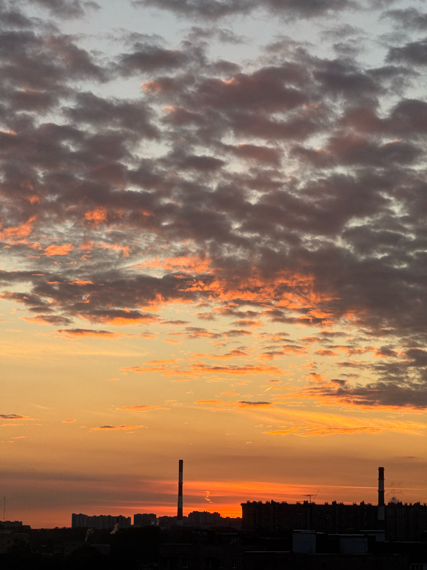 The image depicts a sunset over an urban area, likely Saint Petersburg, Russia. The sky is filled with a dramatic display of clouds, tinged with orange and pink hues from the setting sun, contrasting with darker, grey clouds above. Silhouetted against the vibrant sky are buildings, a tall industrial chimney with red and white stripes, and what appears to be residential blocks. The foreground is dark, obscuring details of the immediate surroundings, suggesting the photograph was taken from an elevated position. The overall scene conveys a sense of twilight and industrial landscape.