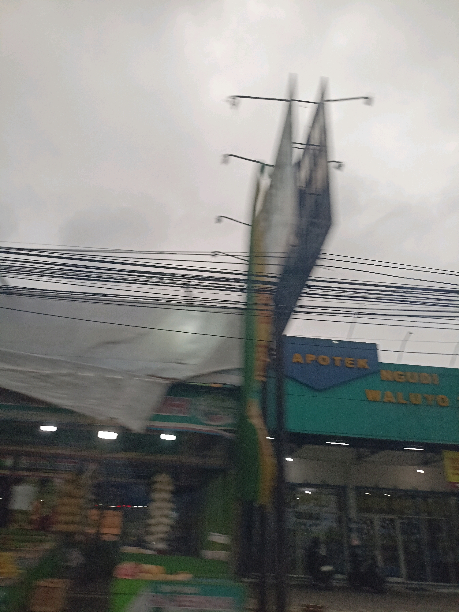 The image captures a street scene in Payaman, Indonesia, characterized by motion blur suggesting movement, possibly from a vehicle. Overhead, a dense network of electrical wires is prominent against an overcast sky. In the middle ground, a green building with a blue triangular sign that reads "APOTEK" (Pharmacy) and yellow lettering "NGUDI WALUYO" is visible. Below this sign, storefronts with glass doors and windows suggest commercial activity. To the left, a makeshift canopy or awning is partially visible, with some goods or displays blurred in the foreground. The overall impression is of a commercial area on a cloudy day, with the blur obscuring finer details of the surroundings and any potential individuals.