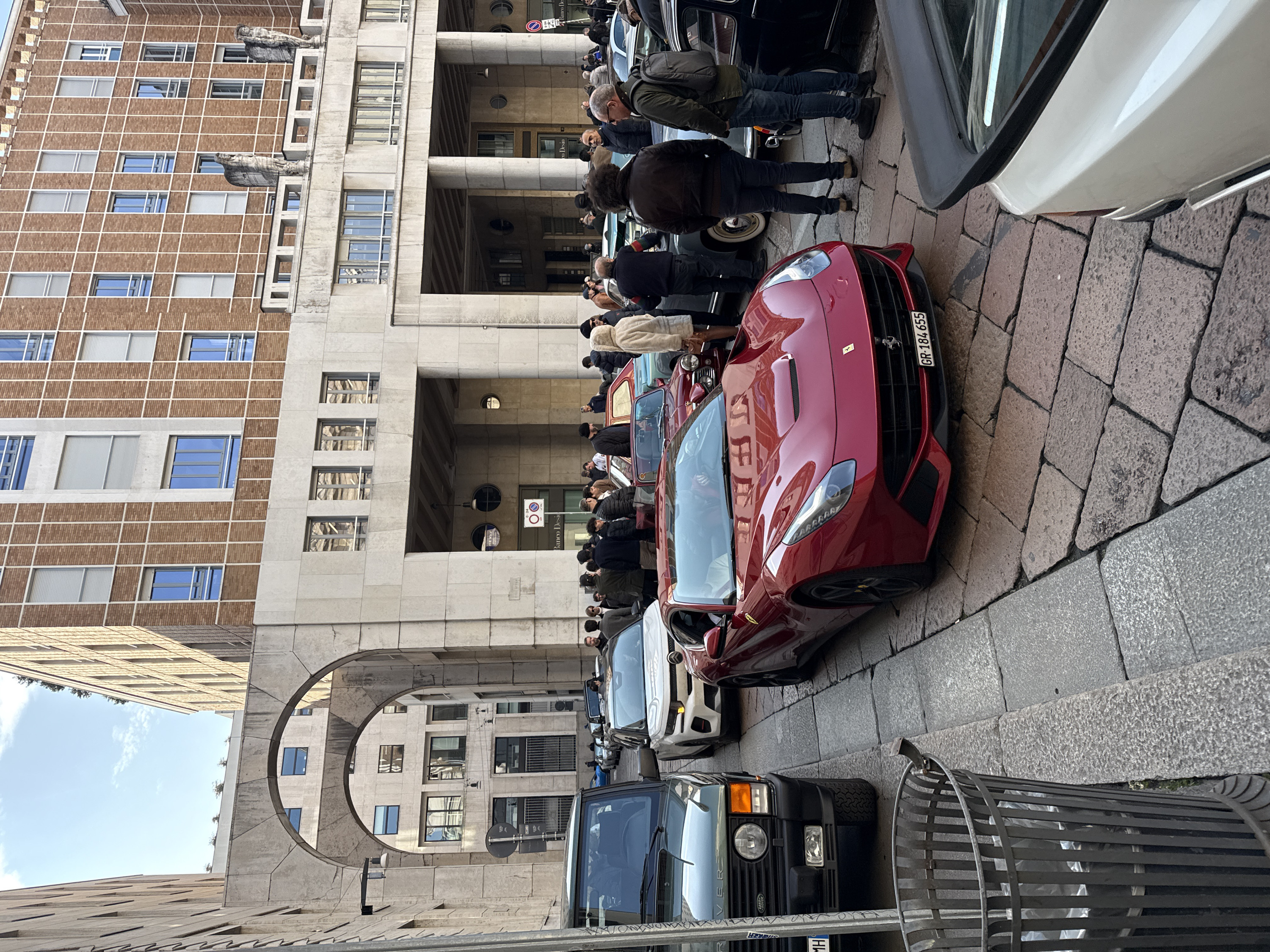 The scene depicts an outdoor urban gathering on a cobblestone street at Piazza Meda, Milan, Italy, under a clear blue sky with sparse clouds.
In the immediate foreground left, a dark grey metal slatted trash can is positioned next to a raised grey stone sidewalk. A dark green classic Land Rover Range Rover is parked on this sidewalk, its front grill, single circular headlight, and square orange indicator visible.
The mid-ground is dominated by a bright red Ferrari F12berlinetta, parked diagonally on the reddish-brown cobblestone street, facing slightly right. Its Italian license plate, "GR 184 655," is clearly displayed. Behind and to the right of the Ferrari, parts of several other vehicles are visible, including a cream/white car with a black rear spoiler, a dark blue classic car, and a classic red Mini convertible. A large crowd of approximately 30-40 individuals, mostly adult males in casual winter attire, are dispersed throughout the mid-ground and background, standing, observing the vehicles, and conversing.
The background features distinctive architecture. To the left, a light-colored stone building presents multiple arched passages, through one of which a building with reflective windows is visible further down the street. In the center and right, a multi-story building with a brown brick facade segmented by a white grid pattern rises prominently. This building features numerous rectangular windows, some with internal blue tinting and some with external white roll-down shutters. Below the brick section, a substantial stone base contains multiple deep archways, some featuring circular windows above them. A "Banco Desio" sign is visible within one of these archways. Stone statues resembling draped figures are situated on a ledge along this building's facade. A red and blue "No Stopping/No Parking" sign is present near one of the arches.