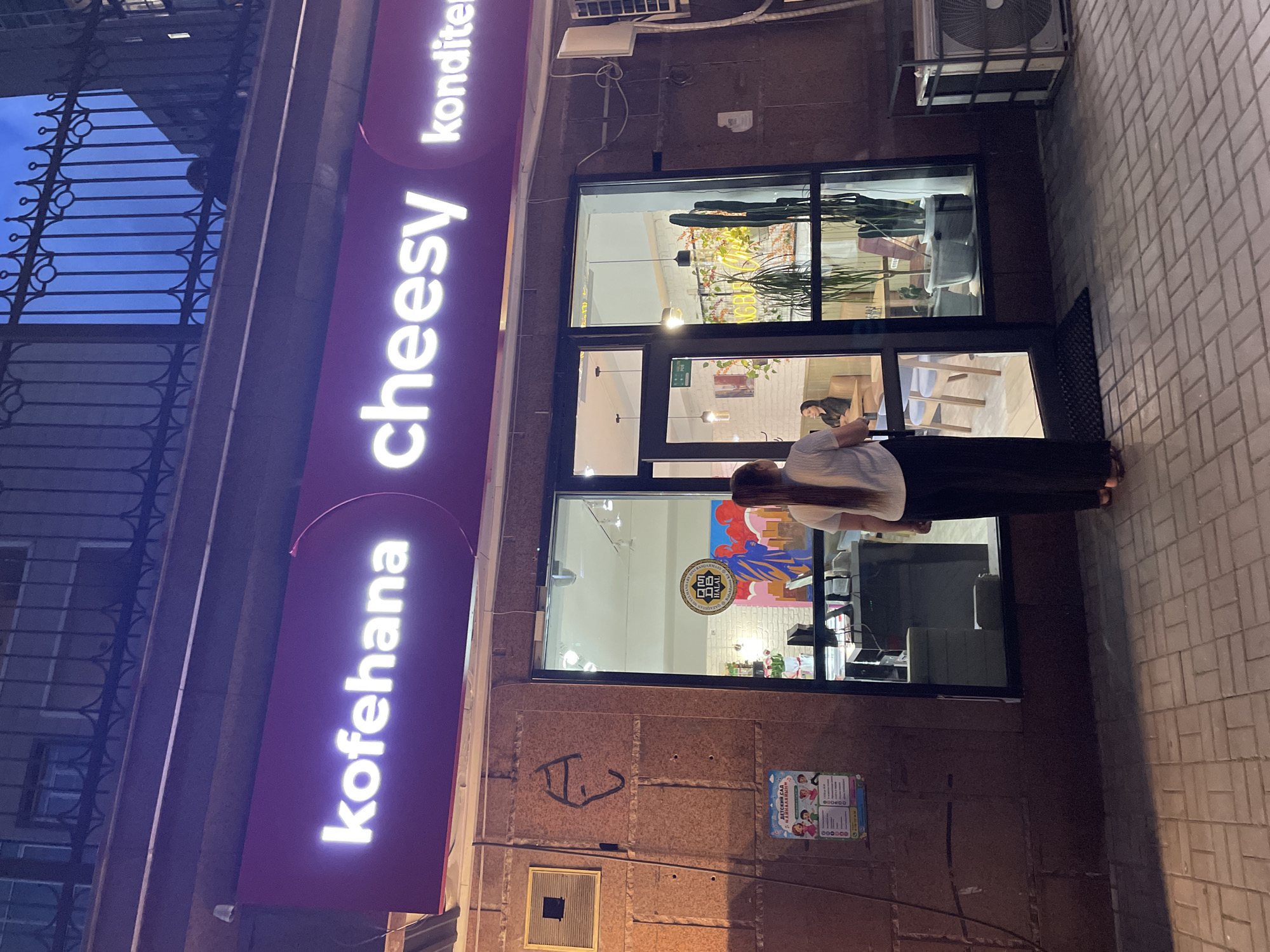 A storefront, likely a cafe, is illuminated by bright white neon signs spelling "kofehana" and "cheesy". A third sign, "konditer", is partially visible. The building exterior is made of rough, reddish-brown stone. A woman with her back to the camera, wearing a white top and black pants, stands at the entrance. Inside the cafe, through large glass windows, two women are seated at tables, and several plants, including a tall cactus, are visible. A poster with cartoon children and text in Cyrillic is displayed on the left wall outside the cafe. The pavement in front of the store is paved with grey rectangular stones. The sky above is dark blue, indicating it is likely evening or night. The overall scene suggests a commercial establishment in an urban setting.