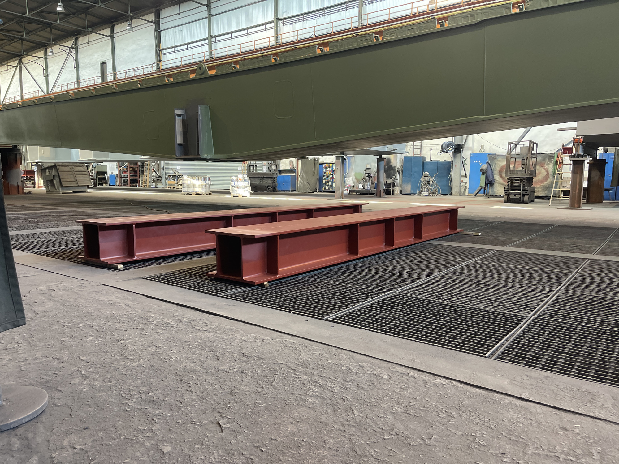 The image shows the interior of an industrial facility. In the foreground are two large, red-primed steel I-beams resting on a grating floor. Overhead, a large, olive-green, box-girder structure spans the space. In the background, various pieces of equipment, tools, and supplies are visible, along with a worker standing near a ladder. The location appears to be a manufacturing or fabrication plant, but the specific city and state are unknown.