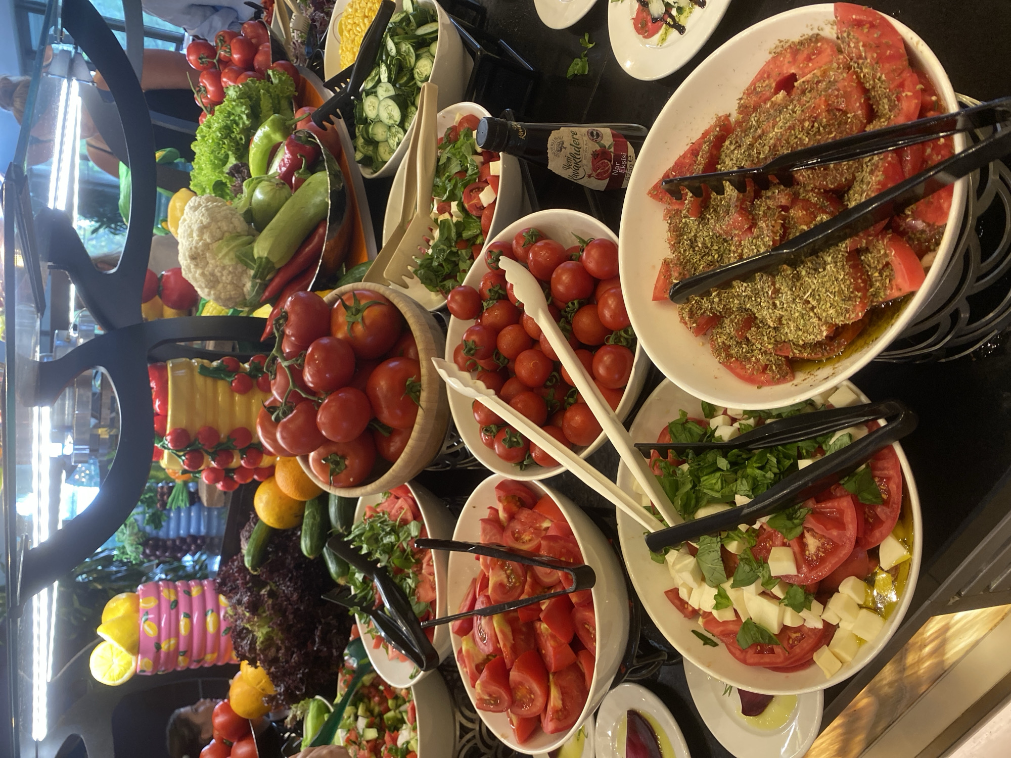 A close-up, eye-level shot captures a buffet display of fresh vegetables and condiments in Aksu, Turkey. The scene is dominated by bowls of vibrant red tomatoes, some sliced and seasoned with herbs, others whole cherry tomatoes, and a salad composed of diced tomatoes, mozzarella cheese, and basil. To the right, a bowl of sliced cucumbers and a separate bowl of corn are visible. In the background, a bottle of pomegranate molasses stands out. Several pairs of tongs are strategically placed within the bowls for serving. The buffet appears to be set up outdoors, possibly at a restaurant or event, with soft lighting illuminating the fresh produce.
