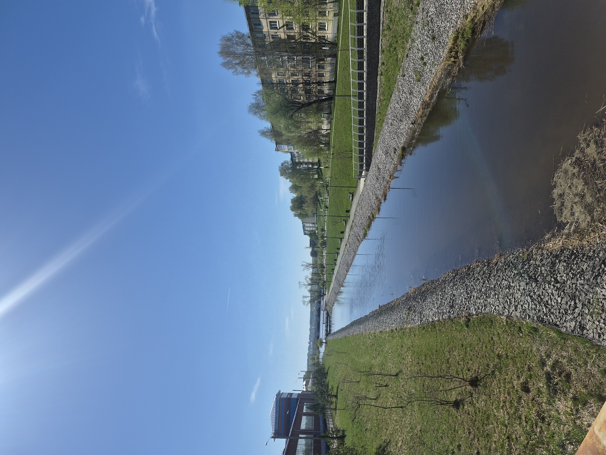 The image shows a canal scene in Votkinsk, Russia, during daylight hours. A canal runs through the center of the frame. Both banks of the canal are reinforced with gray stones and are adjacent to green grassy areas. On the left side of the canal, there's a modern building with a brick facade and blue windows. Streetlights are visible along the grassy area. Across the canal on the right side, there are residential buildings and trees. The sky is blue with some wispy clouds. A bright sun is apparent to the left.