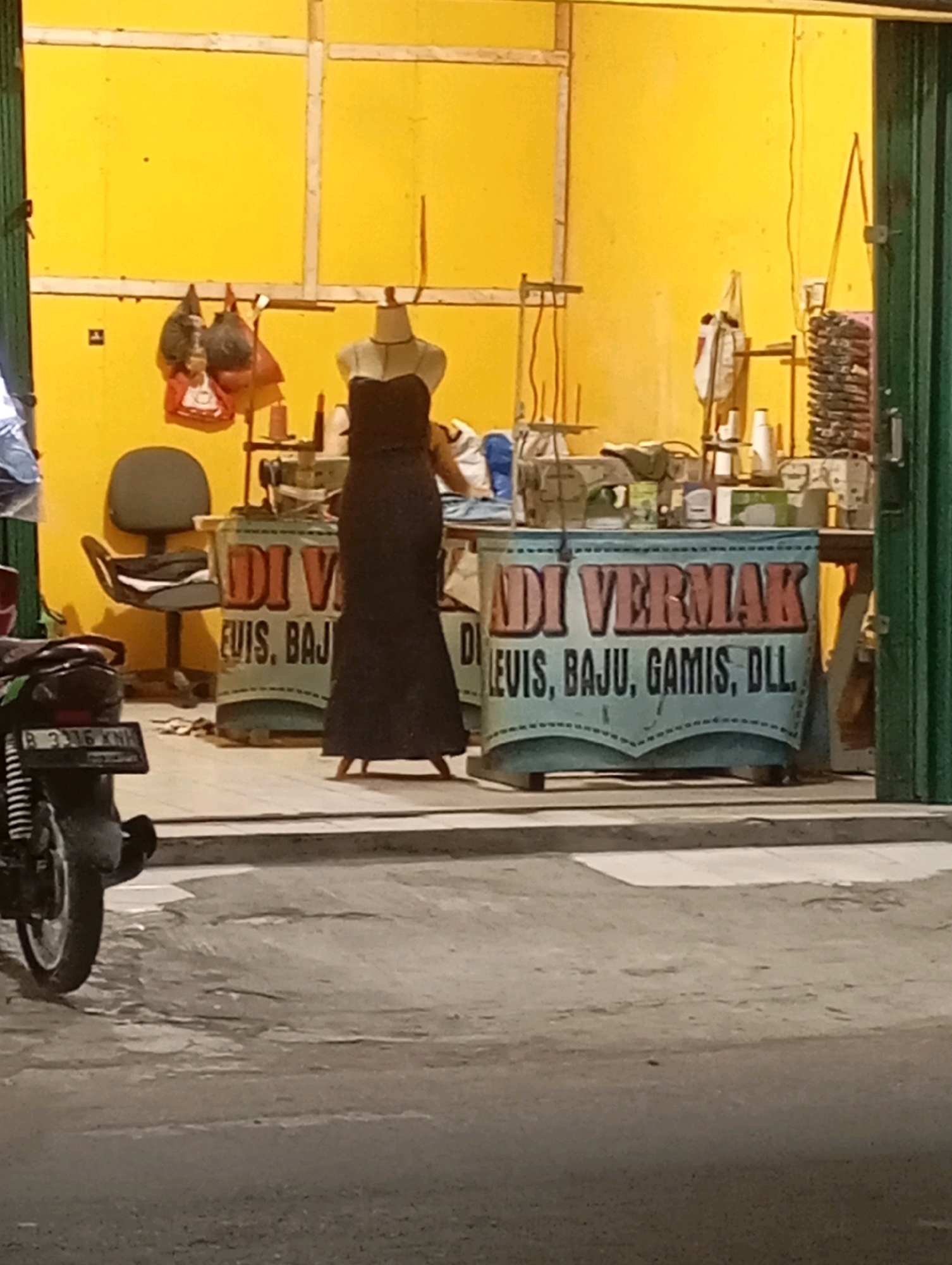 In the city of Babelan, Indonesia, a storefront with a bright yellow facade houses a tailoring and alteration shop. A mannequin dressed in a long, dark gown stands prominently in the center, showcasing the shop's services. In front of the mannequin, a banner displays text in Indonesian, including "ADI VERMAK" and "LEVIS, BAJU, GAMIS, DLL.", indicating that the shop offers alterations for jeans, clothes, dresses, and more. Sewing machines and spools of thread are visible behind the counter, suggesting an active workspace. To the left, a dark-colored motorcycle is parked on the street, its license plate reading "B 3336 KNH". The scene appears to be captured during the evening or at night, with artificial lighting illuminating the storefront.