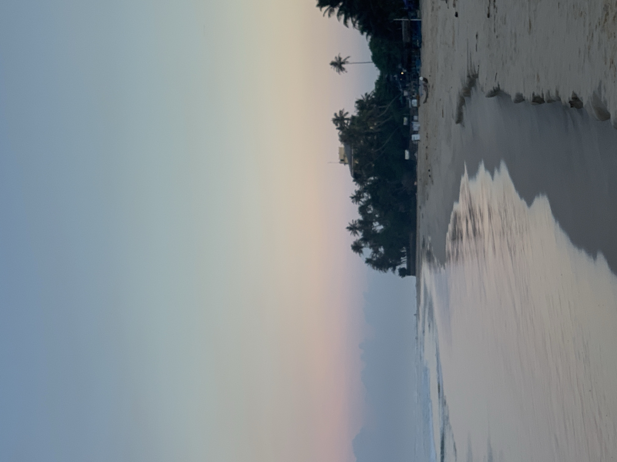 The image depicts a beach scene in Kathaluwa, Sri Lanka. The right side of the frame shows a sandy beach meeting the water's edge. The water reflects the sky, creating a mirror-like effect. Palm trees and other vegetation line the shoreline. The sky transitions from a light blue on the left to a soft yellow and pink near the horizon.