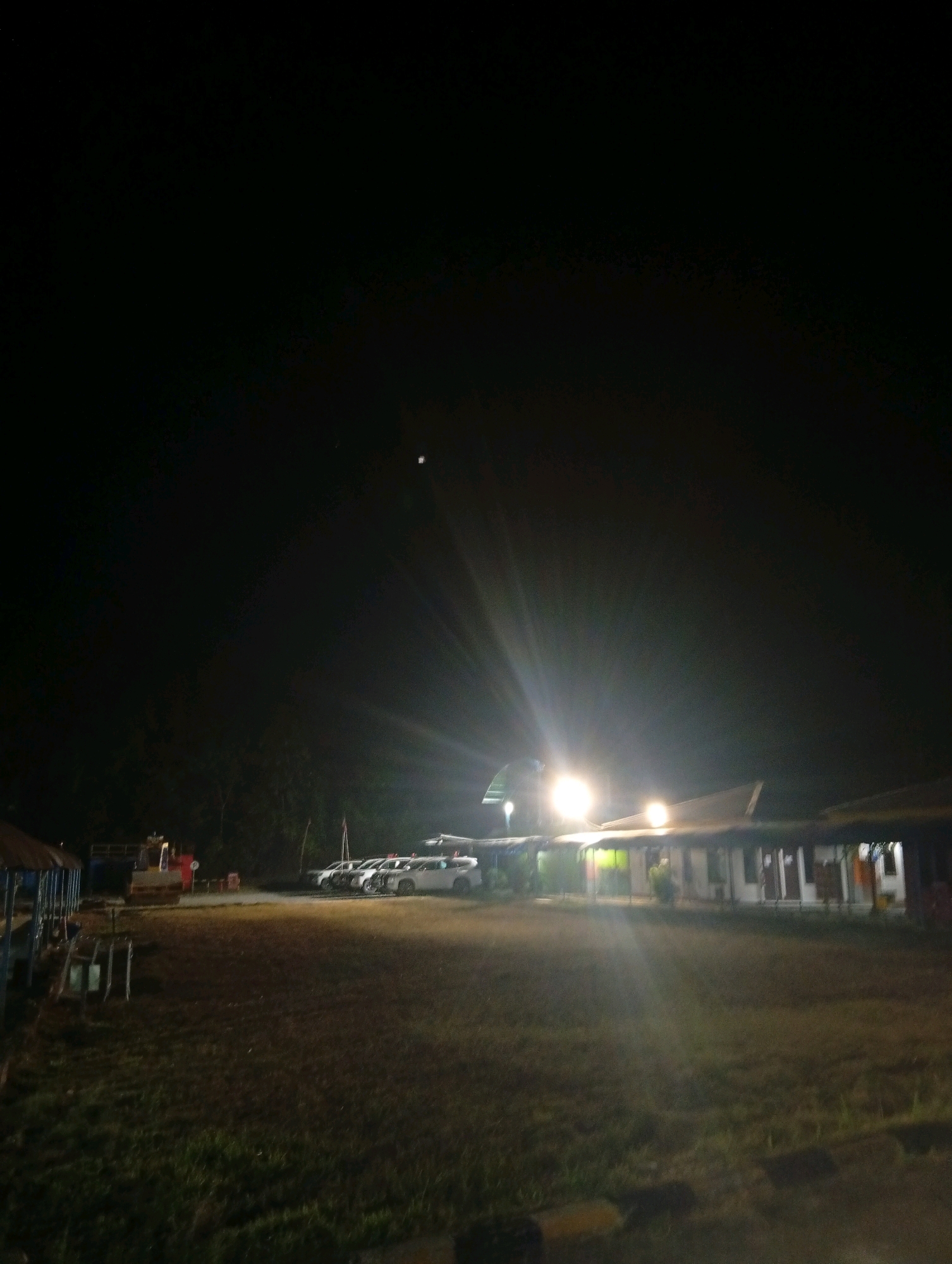 The image depicts an outdoor scene at night in an unspecified location in Indonesia. In the foreground, a grassy area stretches towards a paved or dirt path. Behind this, a building with a covered walkway is visible, illuminated by bright lights that create a glare. Several vehicles are parked in front of the building, some of which appear to be white SUVs or sedans. To the left of the building, there is a structure with a thatched roof supported by blue poles, and a piece of construction equipment, possibly a roller, is parked nearby. The background consists of dark trees and a night sky with a faint hint of moonlight. The overall impression is that of a community area or a small commercial establishment at night.
