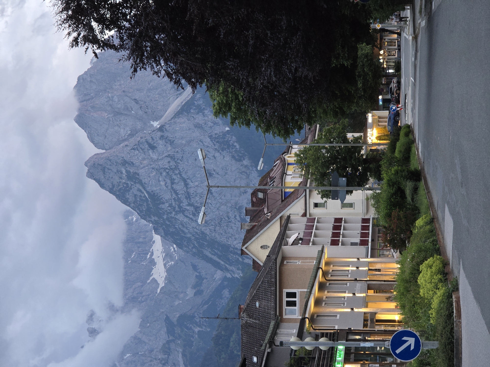 The image depicts a street scene in Garmisch-Partenkirchen, Germany, with a prominent view of the Zugspitze mountain range in the background. The foreground features a paved road curving towards the right, bordered by lush green bushes and sidewalks. On the left side of the road, several multi-story buildings with illuminated windows line the street. These buildings appear to be a mix of shops and residential apartments, some with balconies. A blue circular sign with a white arrow pointing right is visible on a pole.
In the mid-ground, more buildings are visible, including one with a sign that reads "HOTEL". Trees, some with dark foliage, are strategically placed along the street, partially obscuring some of the buildings. The street is relatively empty, with a few cars parked or driving. A solitary figure is walking on the sidewalk in the distance on the right side of the image.
The background is dominated by the majestic Zugspitze mountain, its rugged peaks partially shrouded in dramatic clouds. Snow can be seen on some of the higher slopes, indicating a significant altitude. The sky is overcast, with a mix of gray clouds and patches of lighter sky, suggesting either early morning or late evening. The overall atmosphere is serene, with a blend of urban architecture and imposing natural scenery.