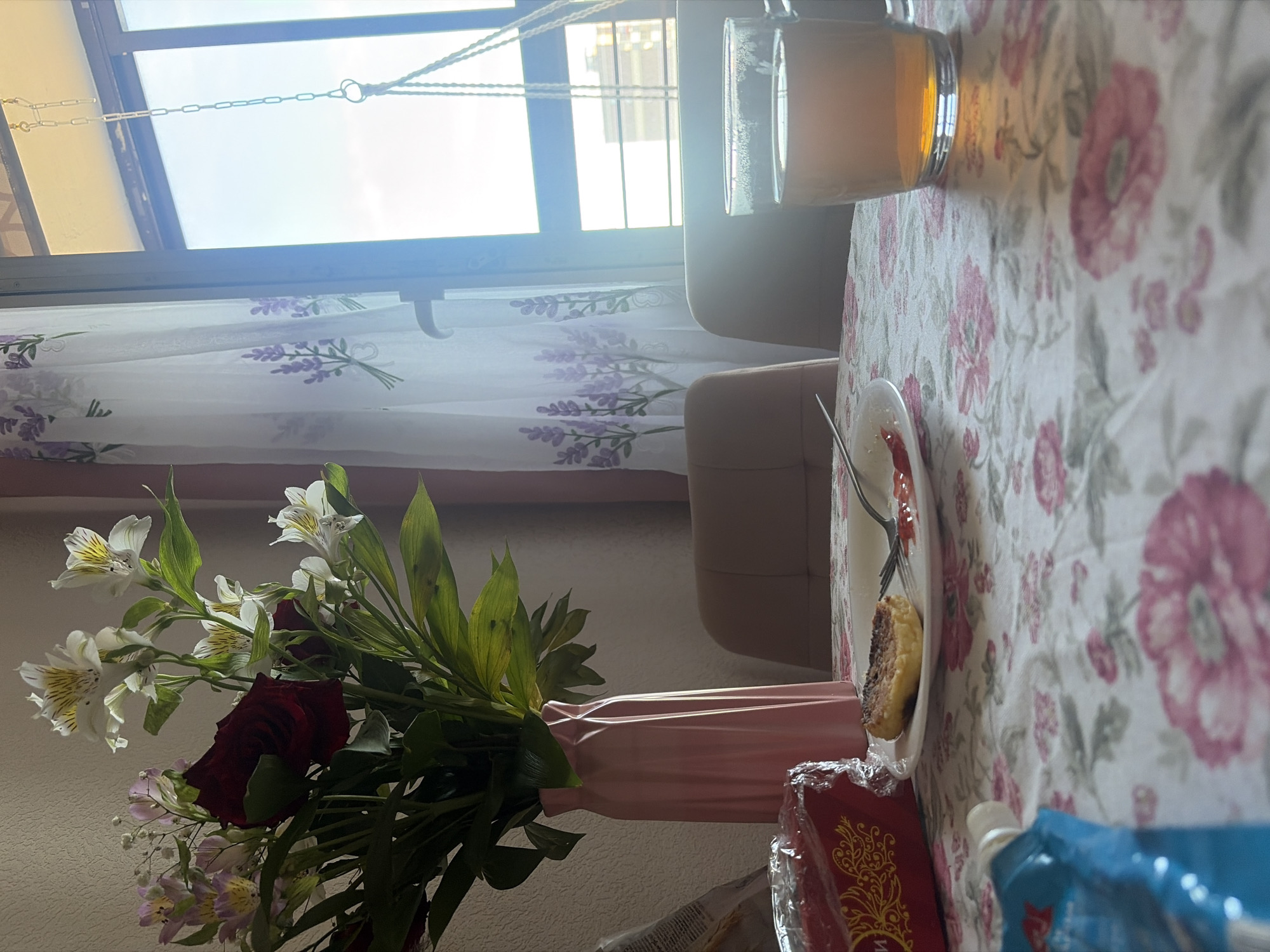 The image depicts a breakfast table in an indoor setting, likely in a home. The table is covered with a floral patterned tablecloth. On the table, there is a plate with food on it, a glass of liquid, a vase of flowers (roses and alstroemeria), and some packaged items. The scene is near a window covered with a sheer curtain with a lavender pattern. Through the window, natural light is visible. The presence of domestic items suggests a residential location. The geographical context, Saint Petersburg, Russia, is not directly visible in the image.