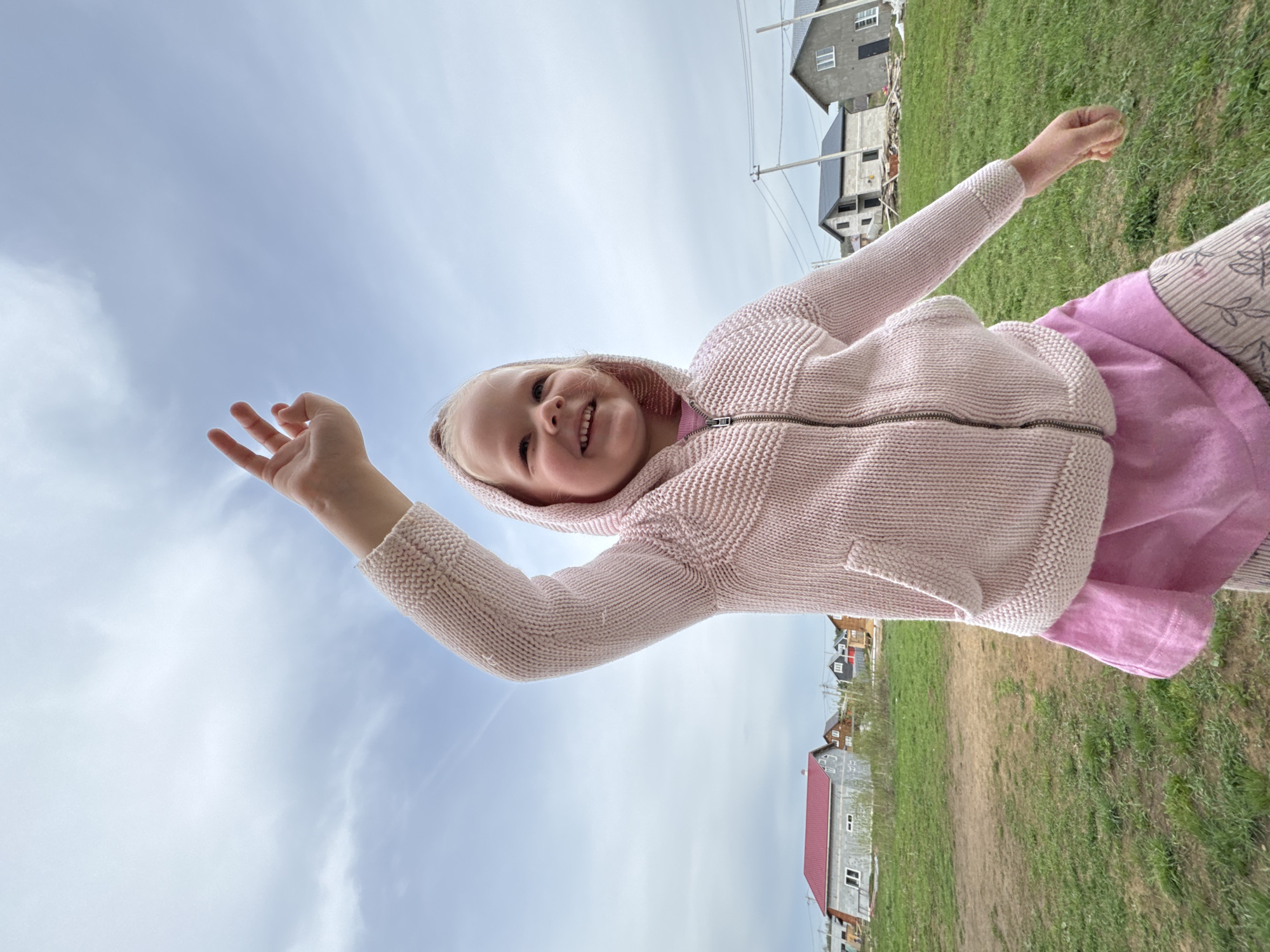 A young girl with blonde hair is facing the camera and smiling in an outdoor setting. She wears a light pink hooded sweater over a pink shirt and patterned pants. Her arms are raised. The background includes a cloudy sky and green grass. A few buildings are visible in the background, along with power lines. Based on the location information provided, this scene takes place in Ферма, Russia.