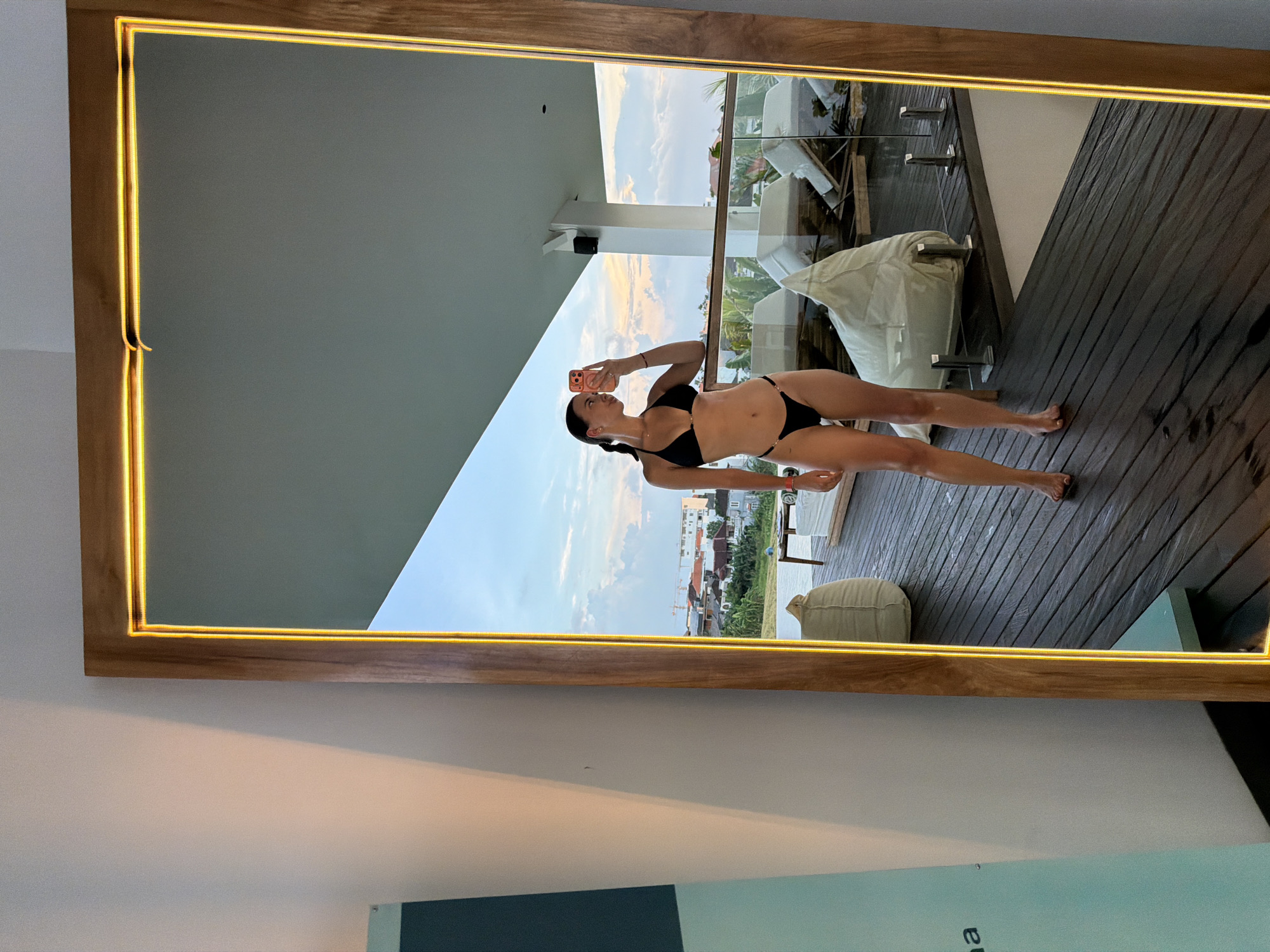 A young woman in a black bikini is reflected in a mirror, taking a selfie with a pink smartphone. She is standing on a wooden deck, likely a balcony, overlooking a city. The city appears to be North Kuta, Indonesia, due to the architectural style and vegetation visible in the background. The deck has white outdoor furniture and a glass railing. The mirror has a wooden frame with a light strip around it. The wall behind the mirror is two-toned, with white at the top and a light blue/green color at the bottom.