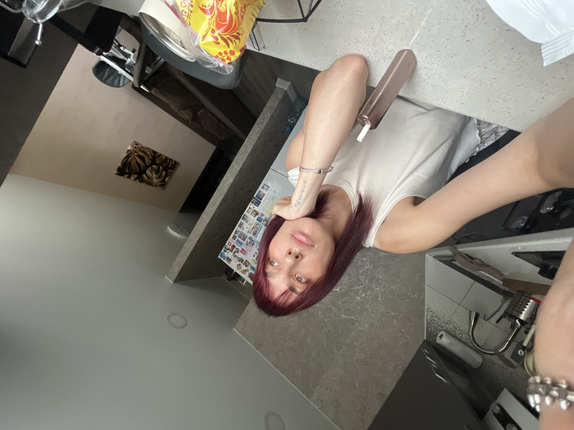 A young woman with dark red hair and bangs is taking a selfie from a high angle, leaning her head on her hand. She is wearing a white tank top and has a tattoo on her left forearm. She is holding a rose gold electronic cigarette in her right hand. She is in a modern kitchen or dining area with gray countertops and cabinets. In the background, a portion of a living room with a couch and a high chair can be seen. The location is identified as Nizhny Novgorod, Russia.