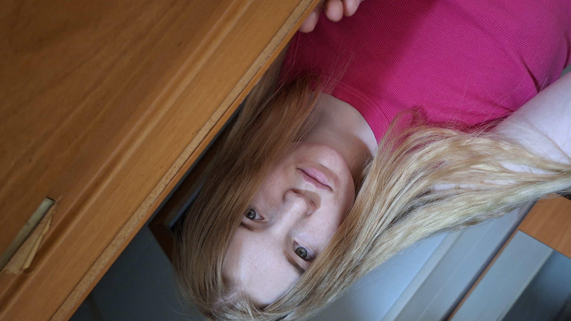 An indoor, rotated portrait features a young woman with fair skin, light blonde hair, and green eyes, looking directly at the camera with a neutral to slightly inquisitive expression. She is wearing a ribbed, bright fuchsia or pink top. Her head and upper torso are visible, with her long, light-colored hair fanning out and partially obscuring the background to her right.
A prominent wooden surface, likely part of furniture like a shelf or table, occupies the upper left and center of the frame. This surface is a light to medium brown color with a distinct wood grain and shows signs of wear, including a small, peeled section on its left edge. The unusual angle of the shot, or its rotation, gives the impression that the woman is looking up from a lower position or peeking from beneath the wooden object.
The setting appears to be a home, suggested by the wooden furniture and soft, even indoor lighting, indicating daytime. No specific time of day or weather can be determined. There is no visible text in the image. The implied location is Большой Букор, Russia.