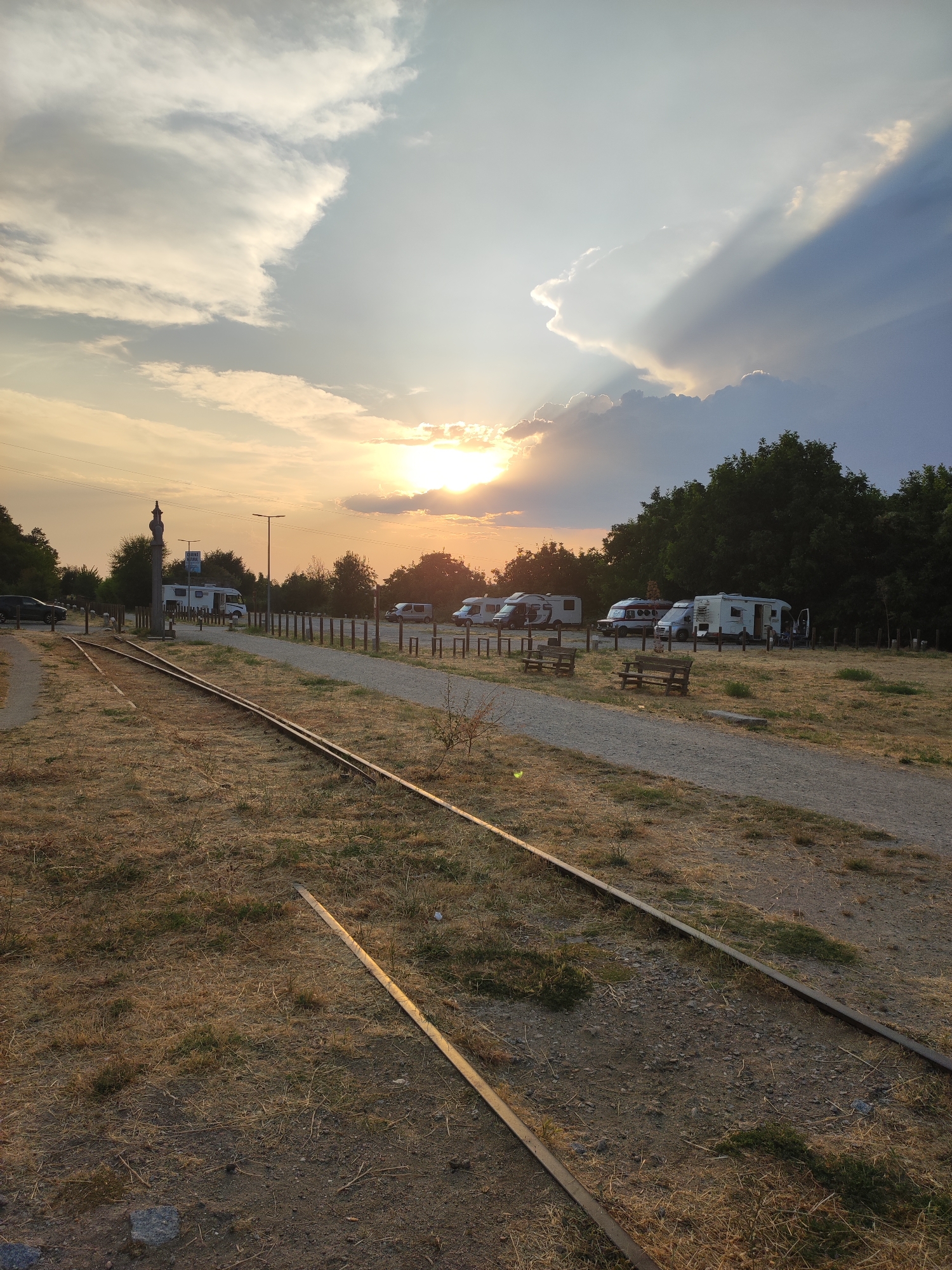 The image depicts a campsite in Béjar, Spain, with several recreational vehicles parked on a gravel lot. In the foreground, disused railway tracks run through dry, grassy terrain. The sun is setting, casting a warm glow and creating dramatic light rays through the clouds in the sky. Trees line the background, forming a dense green backdrop against the sunset. The scene appears peaceful and serene, with no people visible. The overall impression is one of a quiet, natural setting at the end of the day.