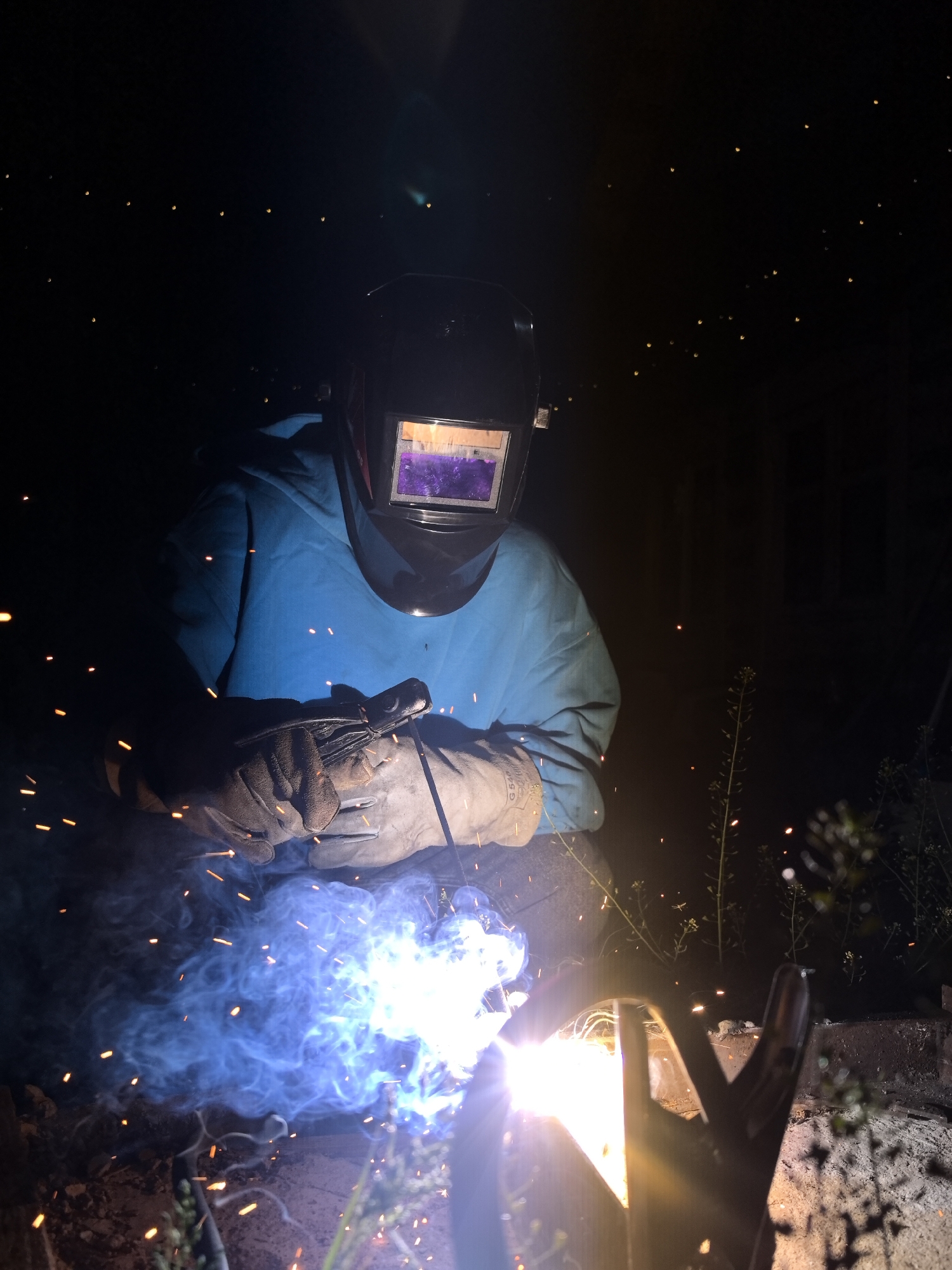 A person is welding outdoors at night. The welder is wearing a black welding helmet with a reflective screen, a blue long-sleeved shirt, and tan gloves. Sparks and blue smoke emanate from the welding area. A metal object is held in place by a clamp in front of the welder. Vegetation is visible in the foreground and background. The location, Noviy, Russia, is not determinable from the visual information available in the photograph.