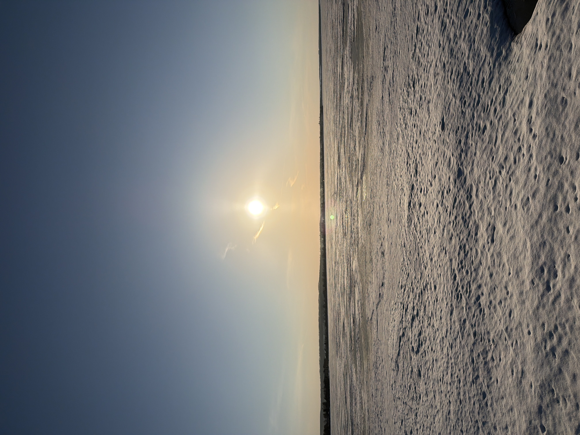 The image depicts a winter landscape in Votkinsk, Russia. The sun is low in the sky, casting a warm glow on the snow-covered ground. The ground is uneven and rough, with many small holes and crevices in the snow. The sky is a clear blue color, with a few wisps of clouds near the sun. In the middle ground, a thin strip of land with trees can be seen. The image appears to be taken from a low angle, making the ground seem to stretch out into the distance. There are no people visible in the scene. The overall impression is one of coldness and desolation, but also of beauty and serenity.
