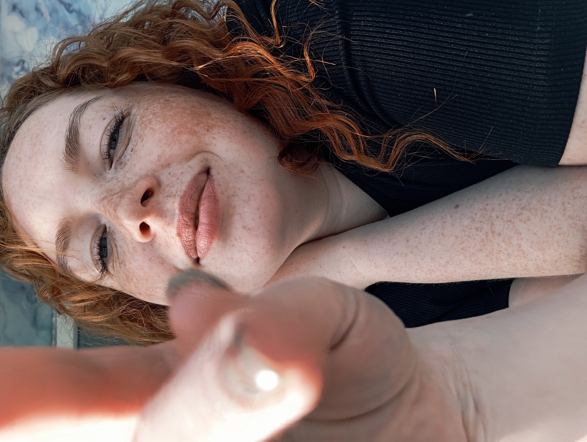 A close-up portrait captures a young woman with vibrant, curly red hair and a scattering of freckles across her face and visible skin. The image is taken from a low angle, giving the impression that she is lying down on her back and looking towards the viewer, with her head positioned on the left side of the frame. Her eyes are partially closed or looking down, and a gentle, relaxed smile plays on her lips, suggesting a soft or contented mood.
She is wearing a black, ribbed top. Her right hand is extended into the foreground, appearing slightly out of focus and blurred, which creates depth and a personal, direct connection with the camera, characteristic of a selfie. Her left arm is also visible, resting on a surface. The background is softly blurred but shows a light, possibly blueish or marbled surface, indicating a bright, possibly outdoor setting during the daytime. The lighting is natural and even, highlighting her features. No visible text is present in the image.