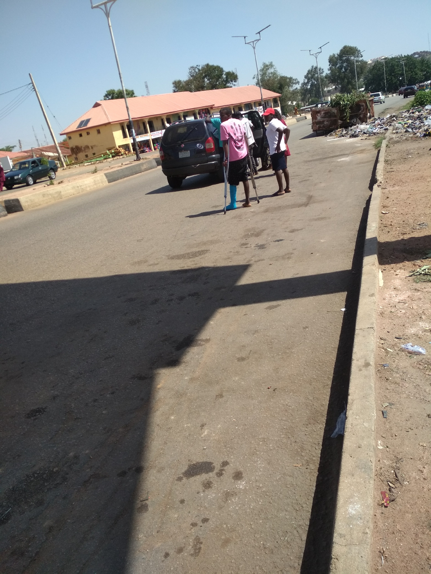 The image depicts a street scene in Gura Top, Nigeria. A man with a cast on his left leg, using crutches, stands on the road near a dark-colored car. Another man in a white shirt and red hat is standing beside him. Behind them, a large container overflowing with trash is visible. In the background, there is a two-story yellow building with a red roof, and other vehicles can be seen in the distance. The road is paved and there is a sidewalk on the right side of the image. The sky is blue and clear.