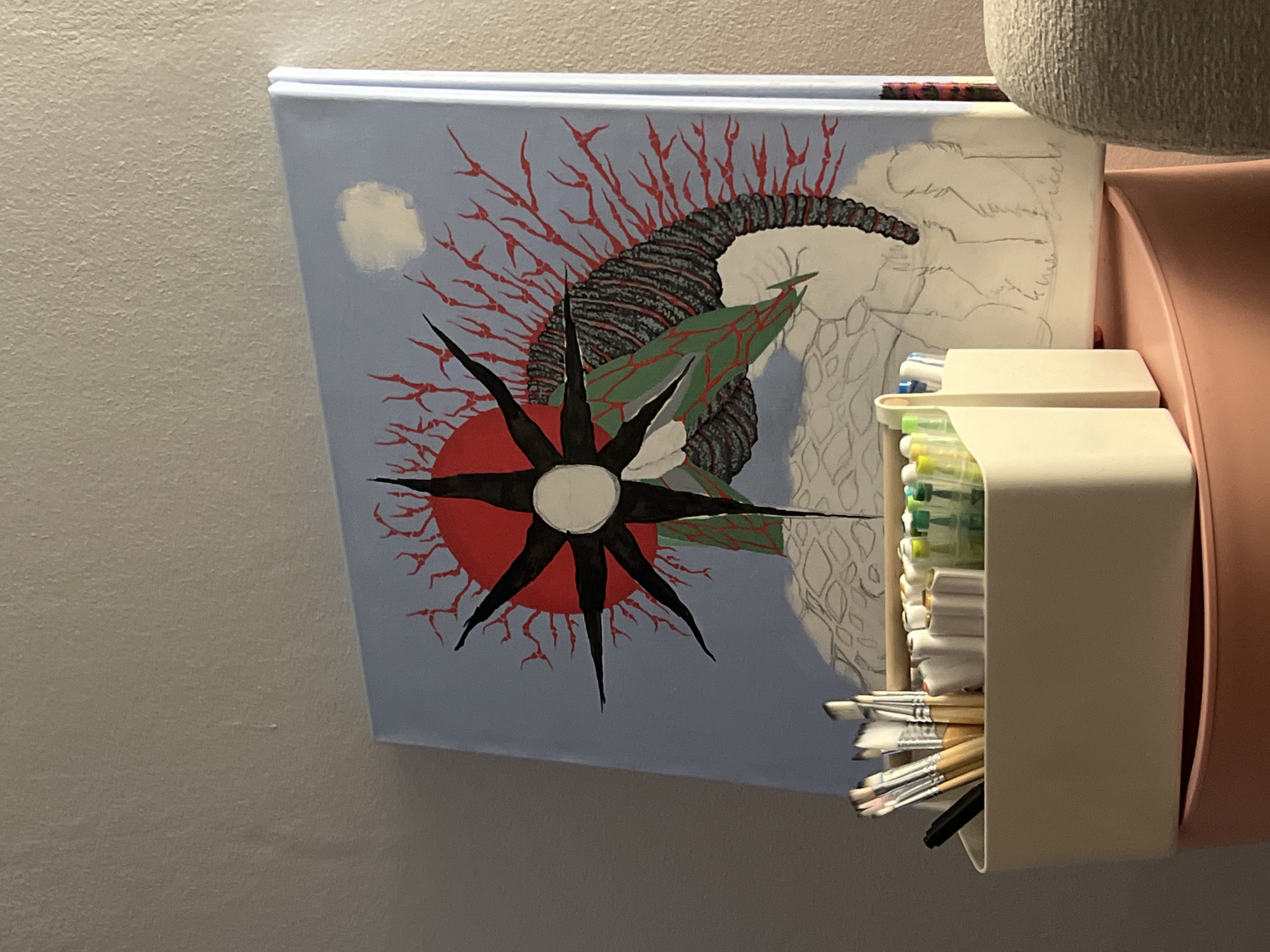 The image shows a close-up view of an artwork on a light blue surface, possibly a canvas or book cover, leaning against a wall. The artwork features a central black starburst with sharp points against a vibrant red circular background. Emerging from behind the starburst is a dark, textured, horn-like shape adorned with red, branching, coral-like elements. Green, leaf-like shapes are also integrated into the composition. To the right of the artwork, a portion of a sketchbook with pencil sketches is visible. In the foreground, two beige plastic organizers are attached to the wall, filled with art supplies. The top organizer holds pens or markers, and the bottom one is packed with various paintbrushes. A pink holder is partially visible behind the organizers. The overall scene suggests an art studio or a creative workspace. The lighting is even, indicating an indoor setting, and there are no clear indicators of time of day or weather. There is no visible text in the image.