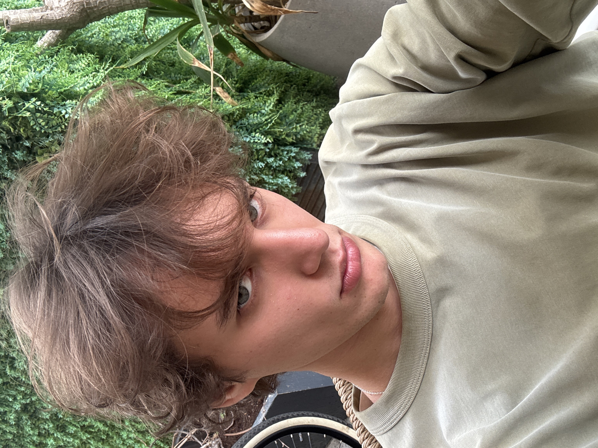 A close-up, angled portrait shows a young man with disheveled brown hair and green eyes. He is wearing a light olive-green crew-neck shirt. The background is filled with dense green foliage, suggesting an outdoor setting. A plant in a gray pot is partially visible at the top. A bicycle wheel is partly visible in the bottom of the frame. While the presence of greenery might suggest a park or garden, there is no indication of the city of Tel-Aviv, Israel.