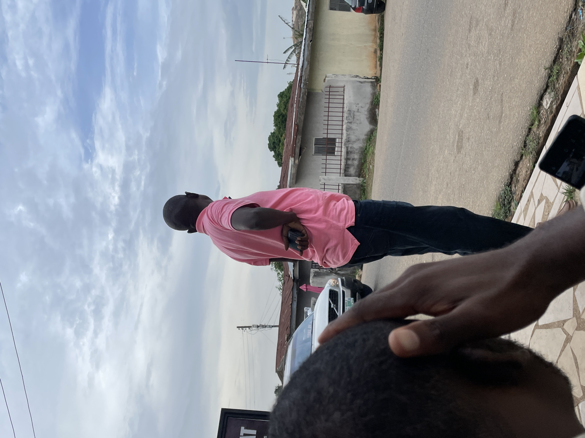 The image depicts a man in a pink polo shirt and dark jeans standing on the side of a paved road. He is viewed from behind, with his head turned to the right, and his left hand tucked into his back pocket. In the foreground, a person's head and hand are visible, partially obscuring the view of the man. A white SUV is parked on the left side of the road, and several buildings with corrugated metal roofs are visible in the background. The sky is cloudy with patches of blue. The scene appears to be in an urban or semi-urban area. The license plate of the white SUV is visible, displaying "KADUNA 515 JNG". This indicates the location is likely in Nigeria, and potentially in the vicinity of Kaduna or another city with a strong connection to that region. Given the context of the prompt requesting details about the city of Jos, Nigeria, it is plausible this scene is situated within Jos, or a related area.