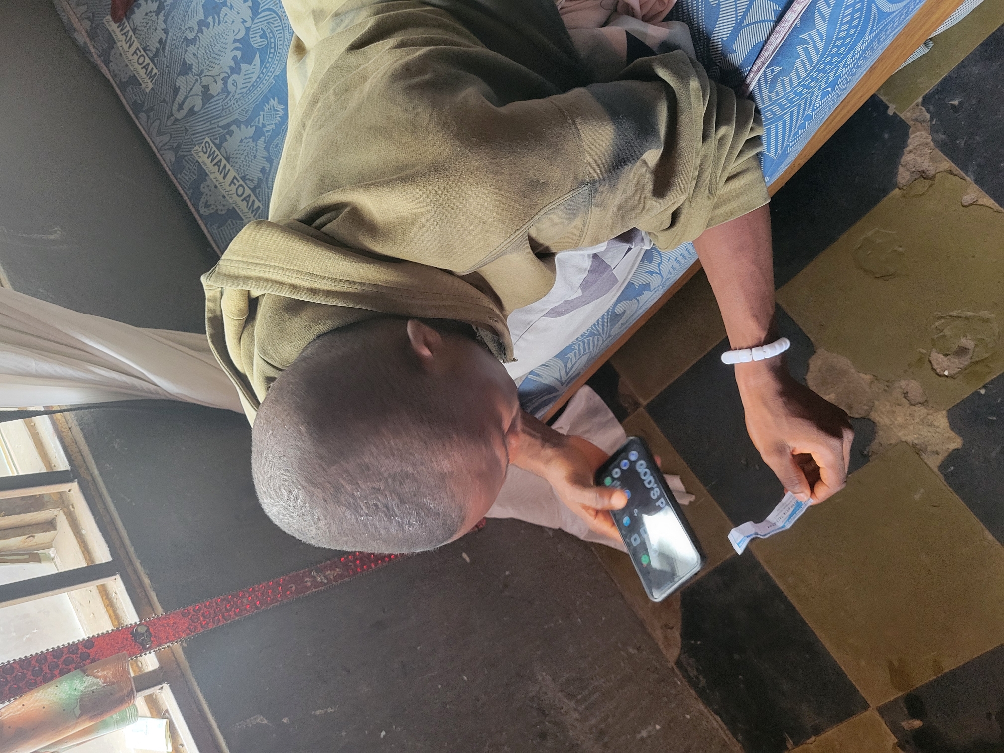A man lies on a "SWAN FOAM" mattress with a patterned blue cover in a room with checkered brown and black tile flooring. He is wearing a tan-colored hooded sweatshirt and a white beaded bracelet. He is holding a smartphone in one hand and a small piece of paper in the other. A window is visible in the lower left corner, and a red belt with decorative skulls is draped nearby. The phone displays the name "GOD'S P..." which is partially obscured. The location is inferred to be in Onitsha, Nigeria.