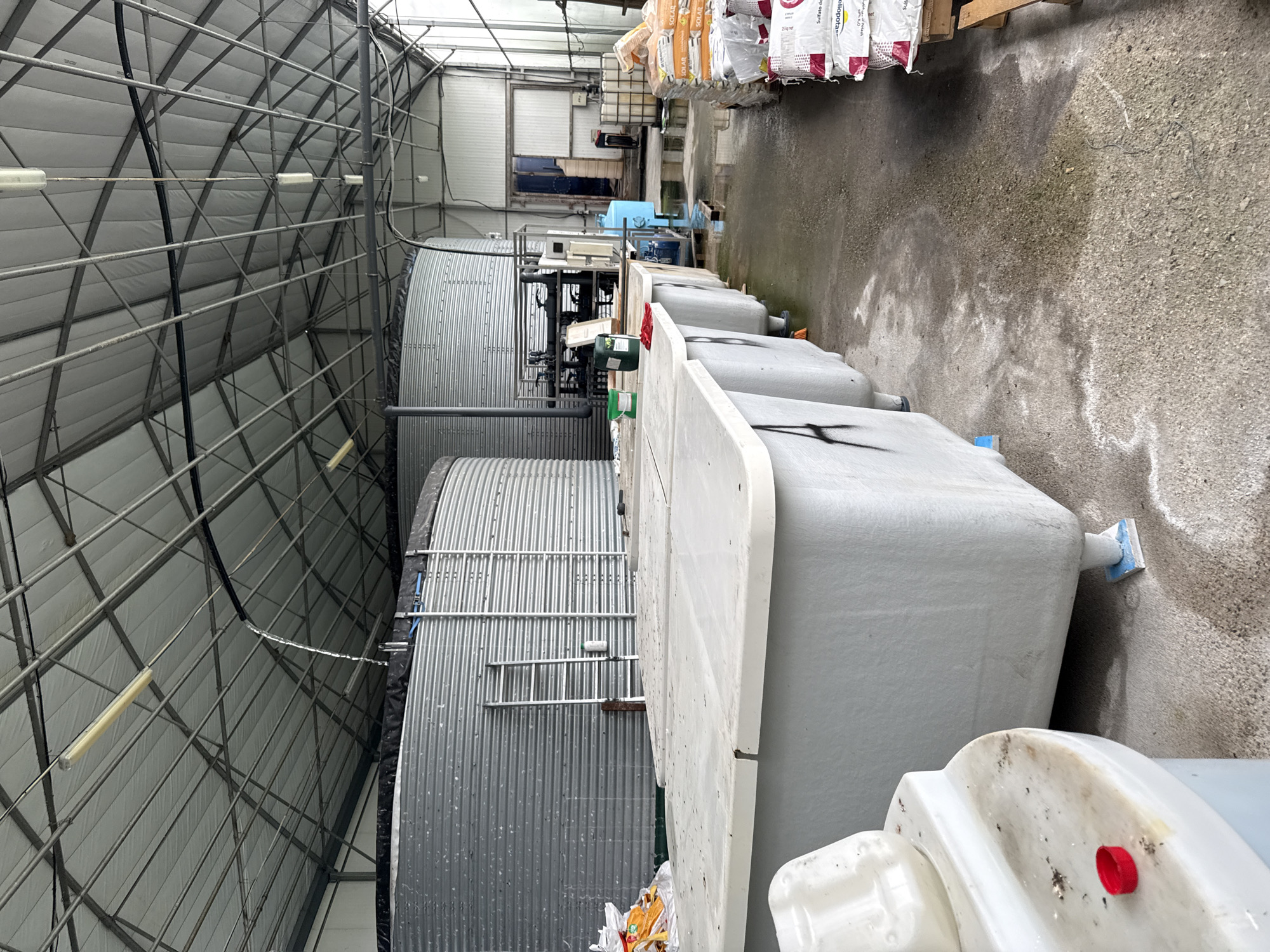 The image shows an interior view of what appears to be an industrial or agricultural facility, possibly located in Stara Zagora, Bulgaria, if that is the provided location context. The scene features large, corrugated metal storage tanks to the left of the frame. Adjacent to the tanks are a series of gray, rectangular plastic containers. Some of these containers are stacked or lined up. Overhead, a metal framework supports the roof of the structure. The floor is made of concrete. Bags are visible in the background.