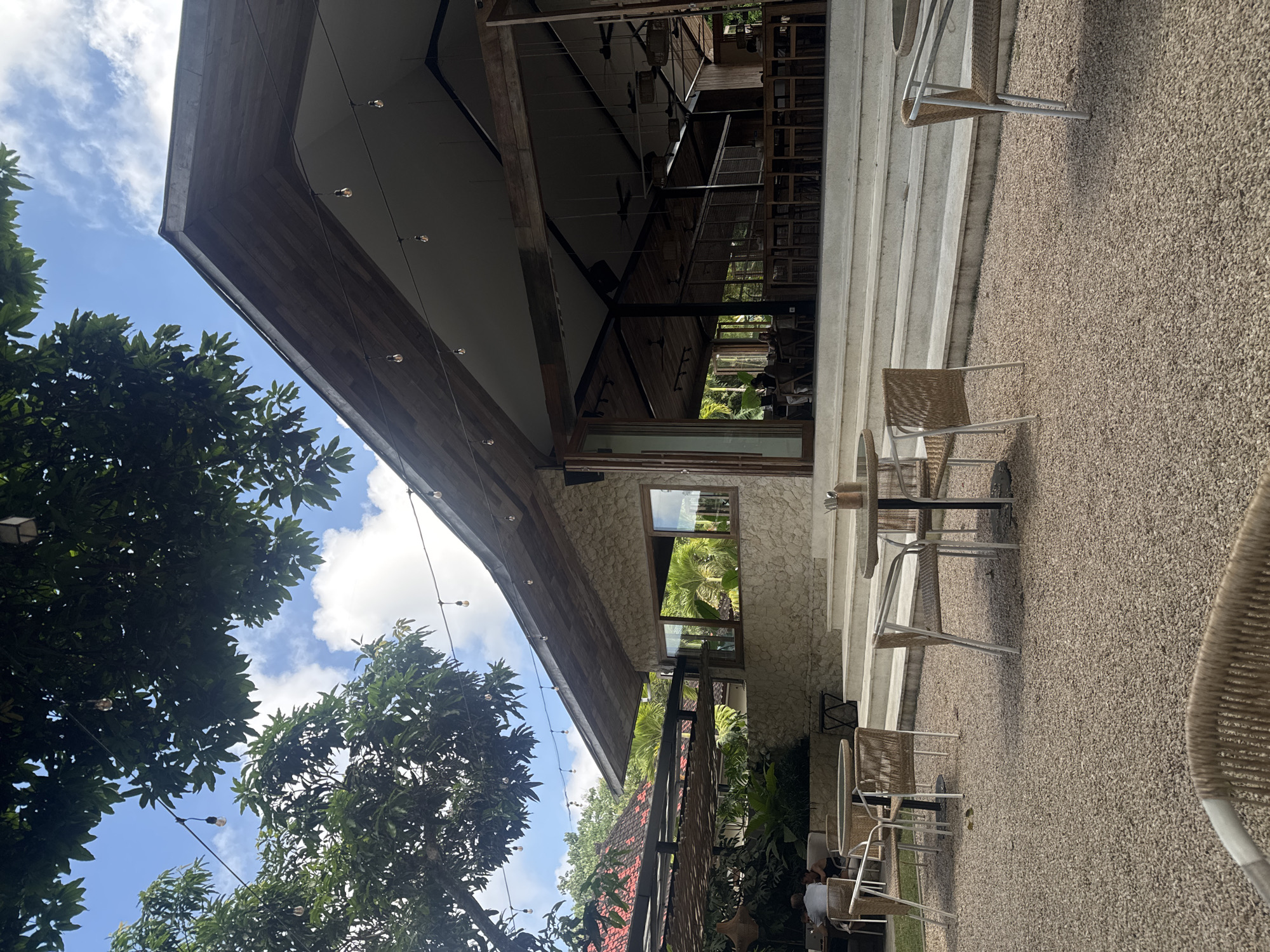 The image shows an outdoor seating area of a building, likely a restaurant or cafe, in Kuta Selatan, Indonesia. The building has a modern architectural design with a wooden roof, a stone exterior, and large windows. There are tables and chairs, seemingly made of wicker, placed on a gravel surface. String lights are visible near the trees in the foreground. The sky is blue with some clouds. People are seated at some of the tables. The scene appears to be taken during the daytime.
