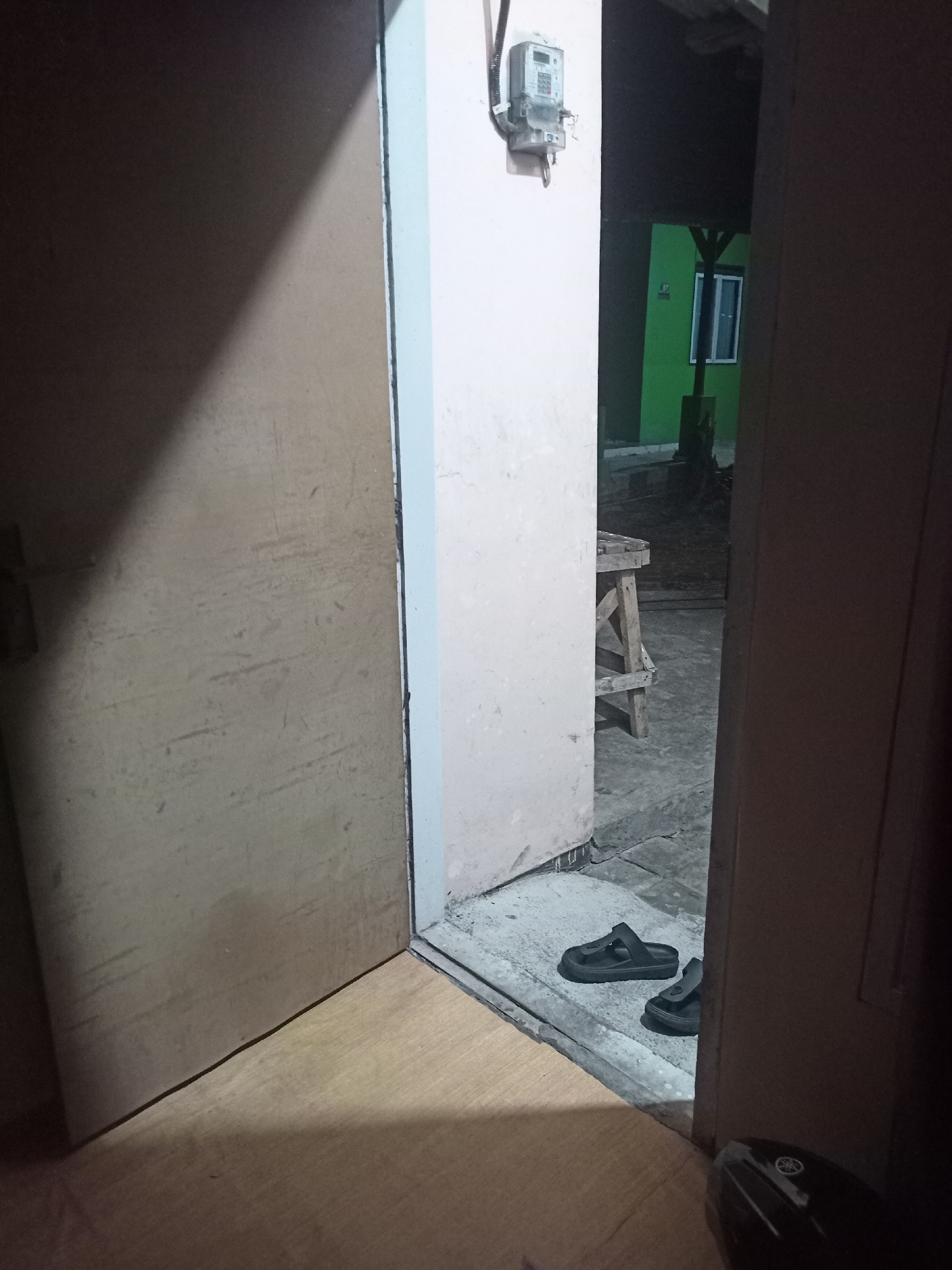 The image depicts an interior doorway area within a residence in Randuagung, Indonesia, during nighttime or low-light conditions. A partially open wooden door is visible on the left, casting a shadow. On the right side, a darker doorway leads to an exterior area where a simple wooden stool is present. A green building with a white-framed window is visible in the background. Black slip-on sandals are placed on the concrete floor near the doorway. An electricity meter is mounted on the pale wall of the interior space. The floor inside appears to be made of light-colored wood, while the exterior ground is paved concrete.