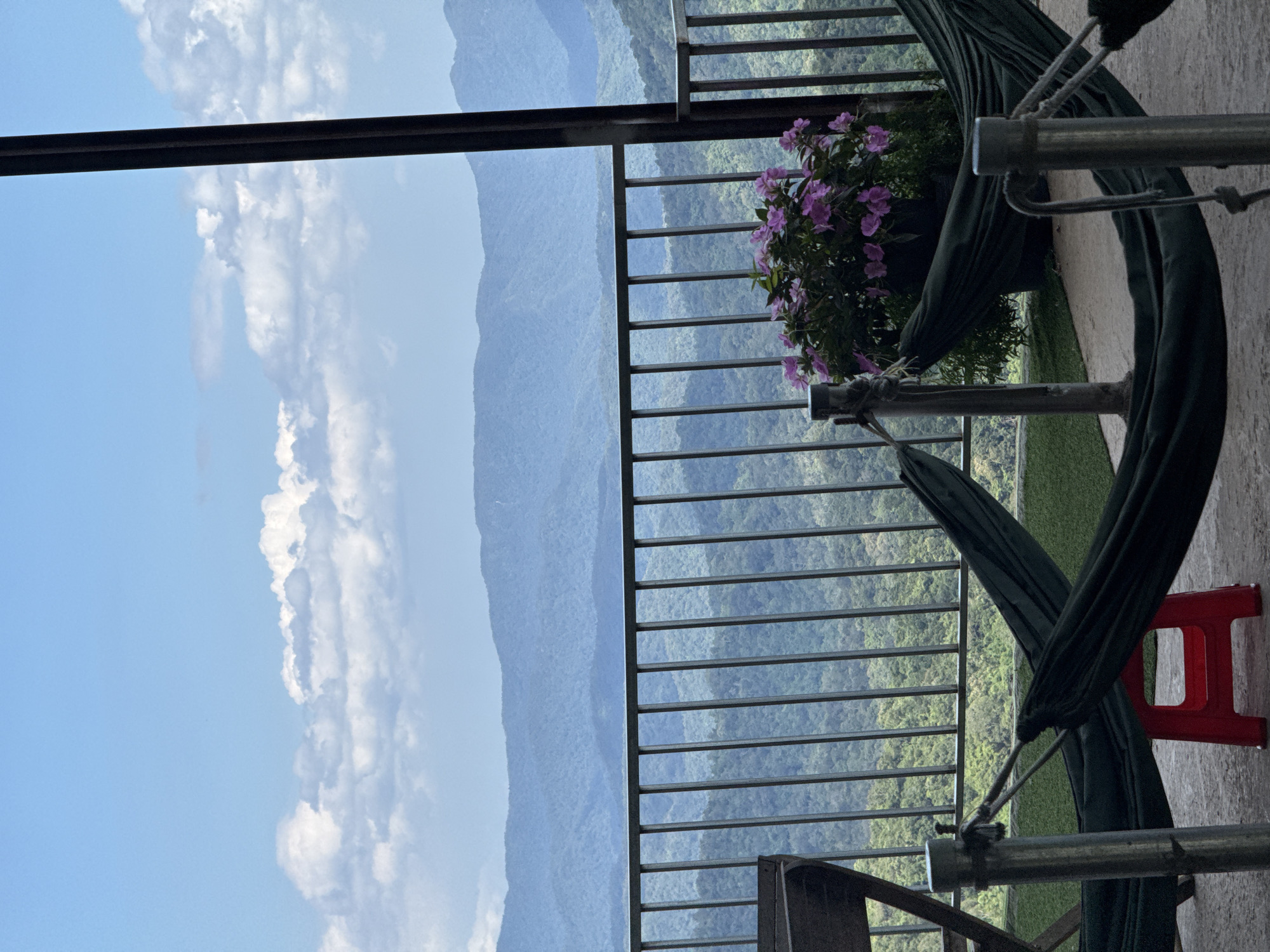 This image depicts a scenic outdoor seating area overlooking a lush, mountainous landscape. In the foreground, two dark green hammocks are suspended between metal posts. To the left, a portion of a wooden chair is visible. In the center, a small red plastic stool rests on the concrete floor. Behind the hammocks, a potted plant with vibrant purple flowers adds a touch of color. A dark metal railing runs horizontally across the scene, separating the seating area from the expansive view of rolling, tree-covered mountains stretching into the distance under a bright blue sky with scattered white clouds. The location appears to be a viewpoint or cafe in or near Phan Son, Vietnam, offering a tranquil escape with a natural vista.