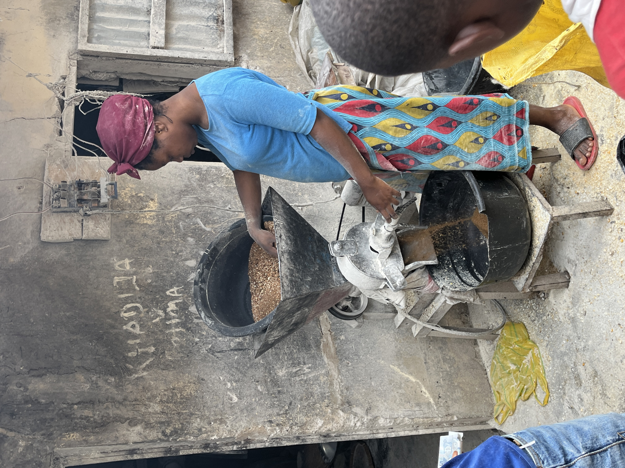 In Bukuru, Nigeria, a woman with a burgundy headscarf and blue shirt operates a manual grain mill outside a building with a rough, dirt-stained concrete wall. The wall features graffiti, including the names "KHADIJA" and "HIMA". She is feeding grain into the top hopper of the mill, which is positioned on a wooden stand. Below the mill, a large black plastic bucket collects the processed grain. To the left of the woman, a yellow plastic bag lies on the ground, and to the right, the back of a child's head is visible, suggesting they are observing the process. The ground is covered with scattered grain. The environment appears to be an outdoor workspace, possibly a market or a small business.