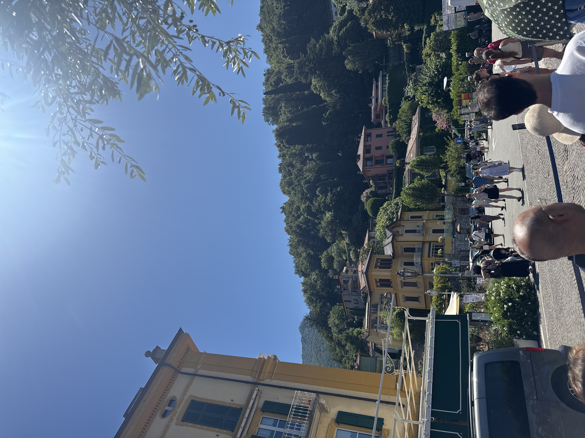 The image depicts a sunny day in Varenna, Italy. In the foreground, a person's head and shoulder are visible, looking towards the scene. The main focus is a street lined with buildings and lush green trees that climb a hillside. Several people are walking along the street, some appearing to be tourists. Buildings with colorful facades, balconies, and shutters are prominent. A yellow building with green shutters is visible in the lower left corner. Scaffolding suggests ongoing construction or renovation on one of the buildings. The sky is a clear blue, indicating fair weather. A large green sign with white text is partially visible, and another sign appears to have Italian text. The overall impression is a lively summer day in a picturesque Italian town.