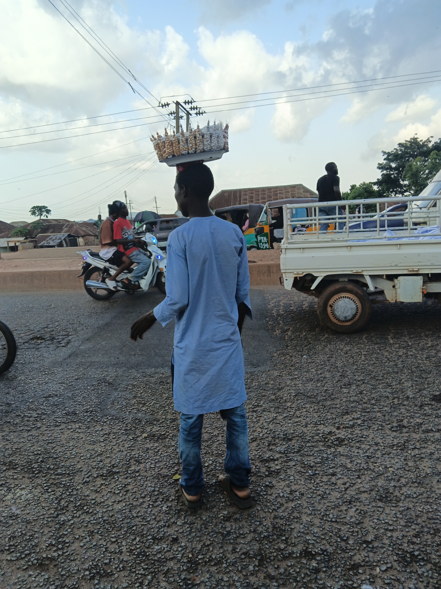In Bukuru, Nigeria, a person is walking on a road, carrying a basket of goods on their head. The person is wearing a light blue long-sleeved shirt and blue jeans. To their left, a white truck is parked. In the background, there are buildings with corrugated metal roofs, a motorcycle with two riders, and other vehicles. The sky is cloudy. The road appears to be paved with asphalt and gravel.
