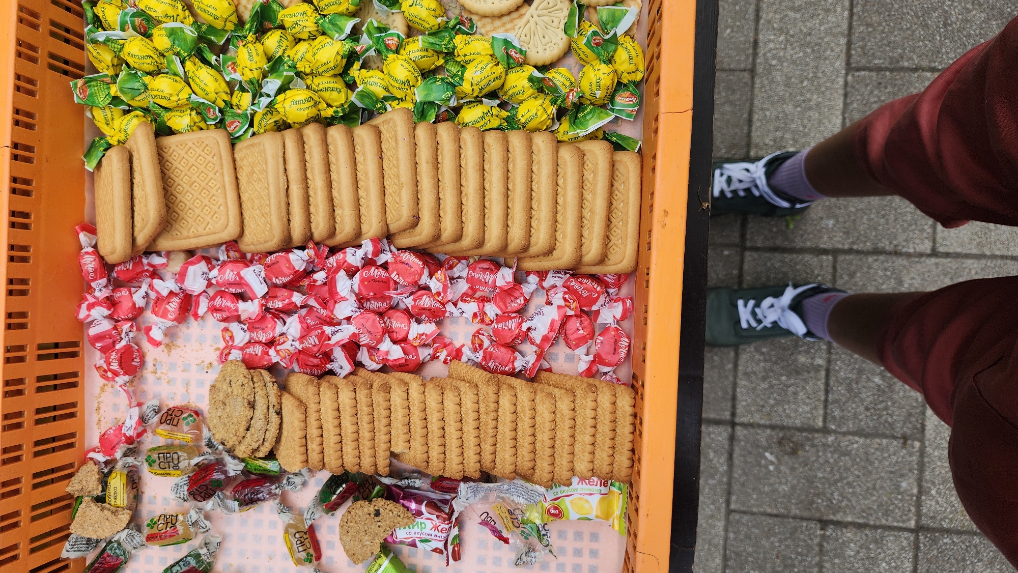 A top-down shot shows an orange plastic crate containing an assortment of sweets and biscuits/cookies. The crate is divided into sections with yellow wrapped lemon candies at the top, followed by rows of rectangular biscuits. Below are red-wrapped candies, and then a section with oat-based cookies and other candies with green and clear wrappers. A person wearing red shorts and green sneakers with white laces is standing next to the crate on a grey paved surface. Some text labels on wrappers appear to be in Russian.